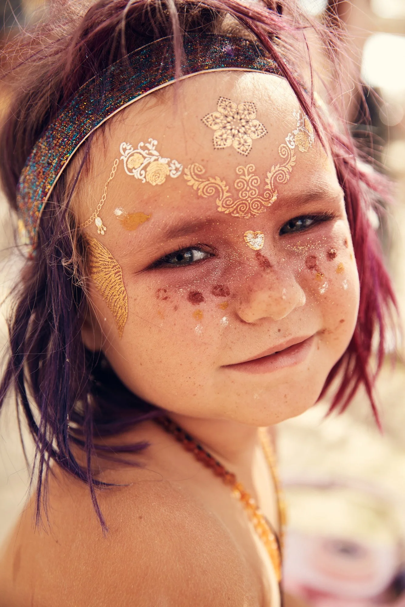Young child with pink hair, glitter, and decorative gold face paint, wearing a sparkly headband at Burning Man in Black Rock Desert Nevada