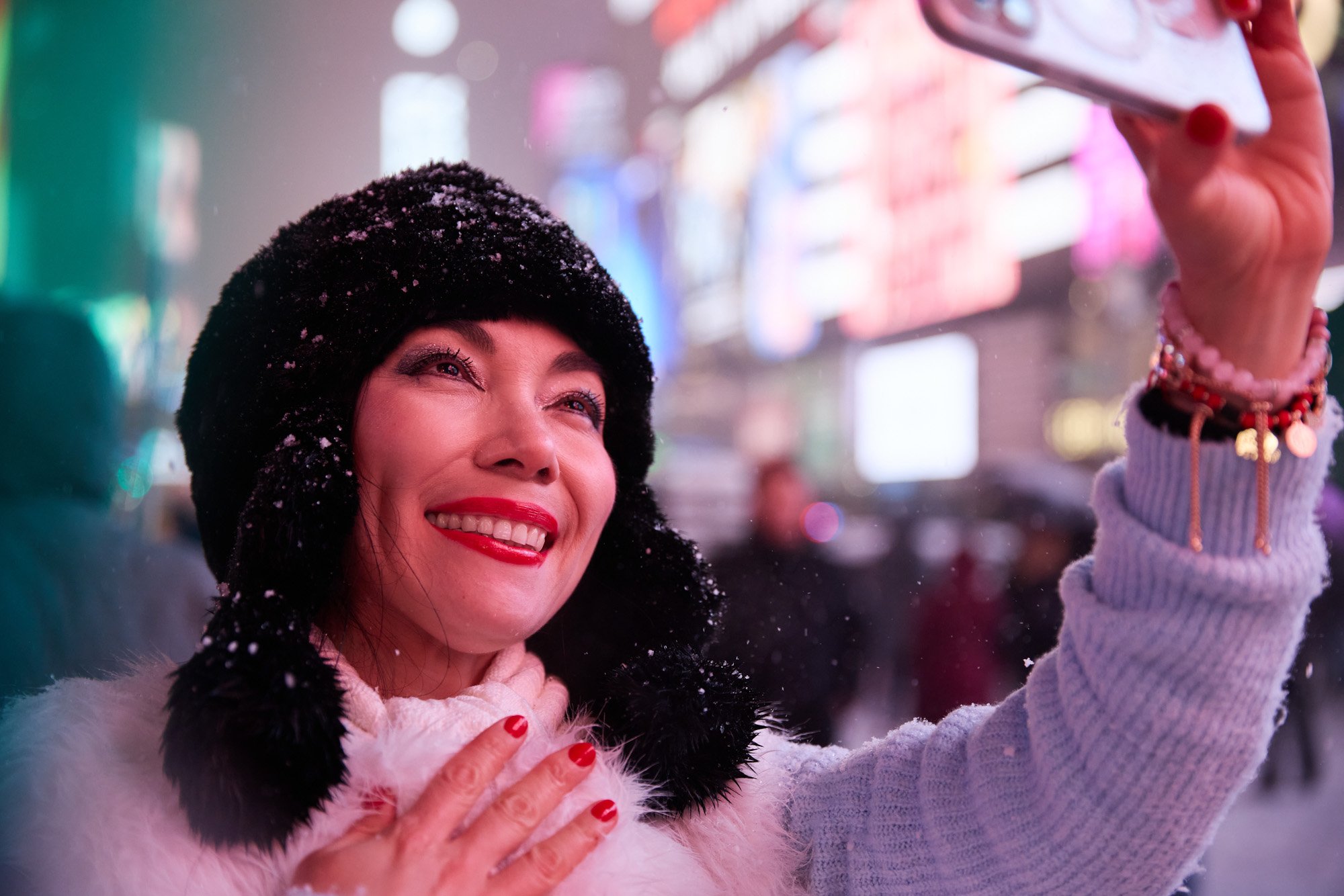 A woman with red lipstick and a smile, taking a selfie in Times Square New York City at night, wearing a black furry hat, a light sweater, and jewelry.