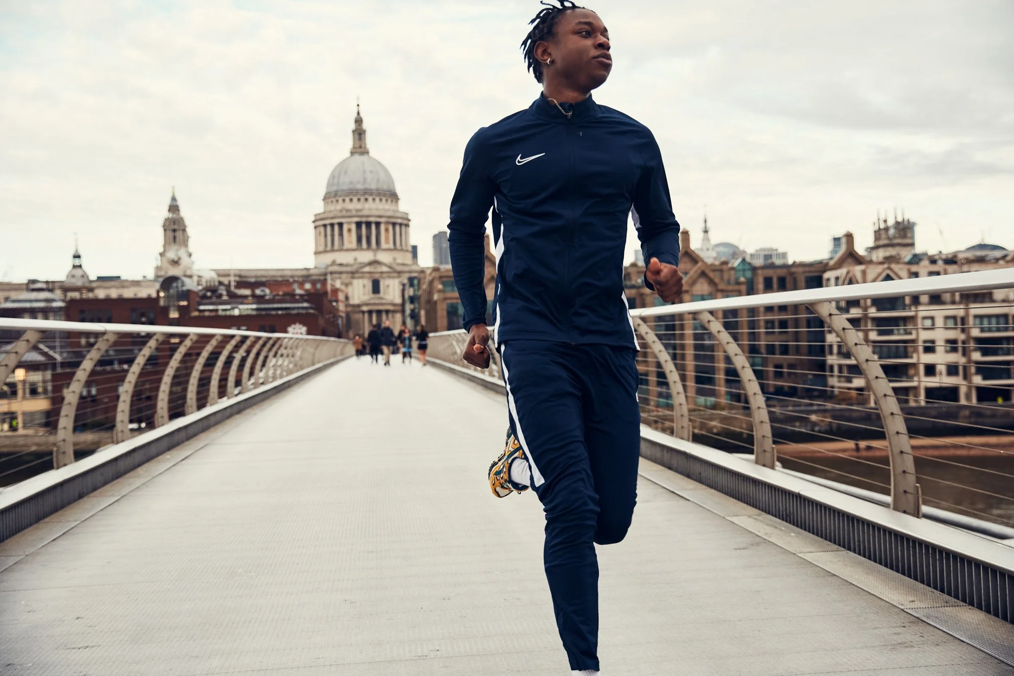 A young Black man jogging on the Millennium Bridge over the Thames River in London, with historic buildings and a domed structure in the background.