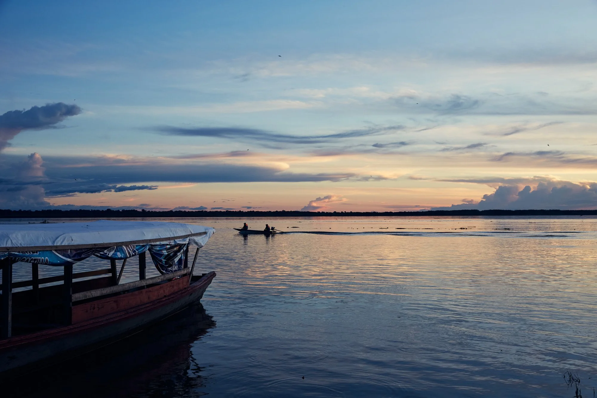 A boat docked at the shore of the Amazon River during sunset in Puerto Nariño, Colombia, with two small boats with people in the distance on the water, and a partly cloudy sky with the sun setting on the horizon.