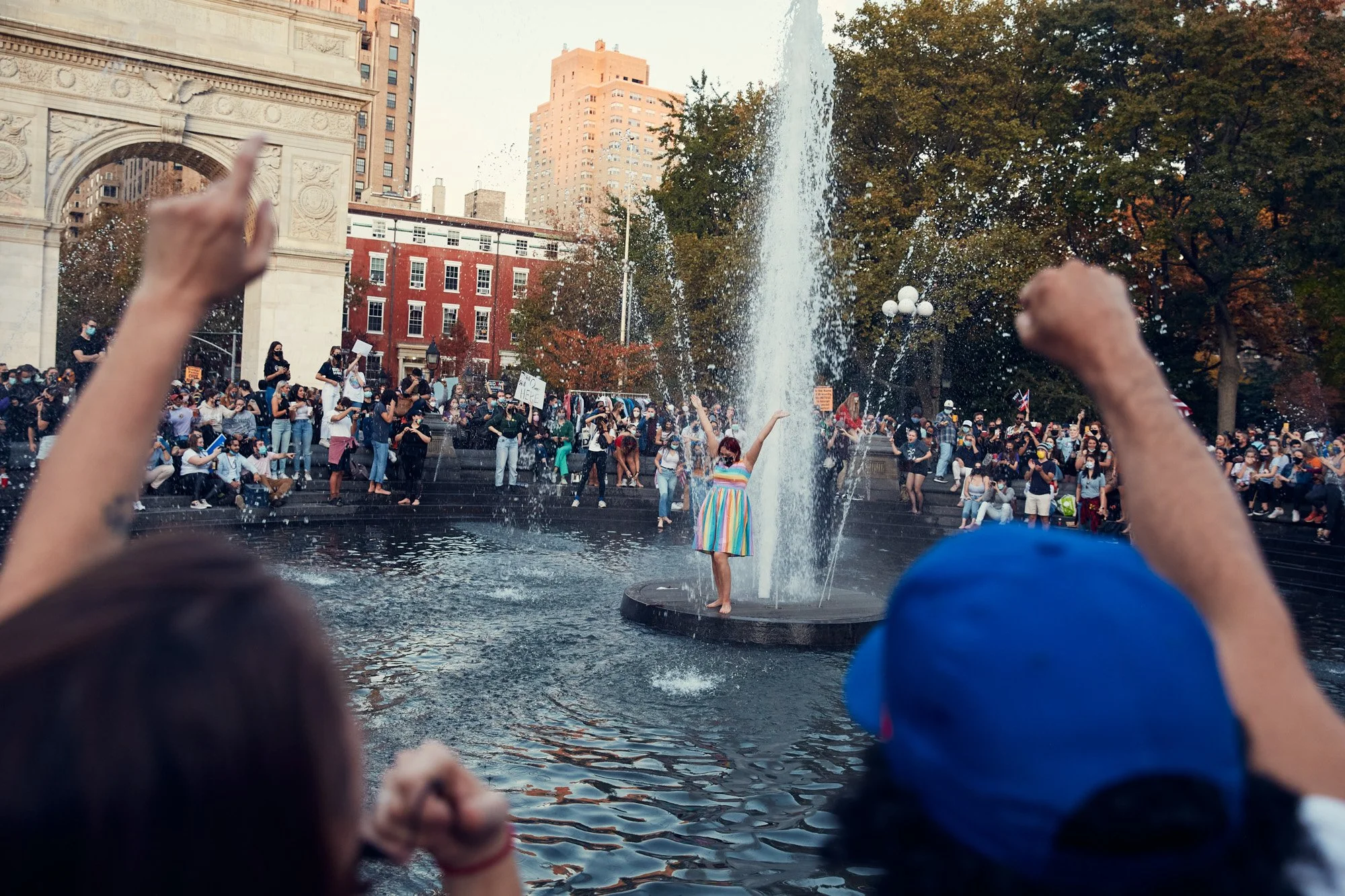 Crowd of people gathered around the fountain in Washington Square Park in Manhattan New York City, with a woman in a rainbow-colored dress dancing in the water, during the daytime. Biden's election day