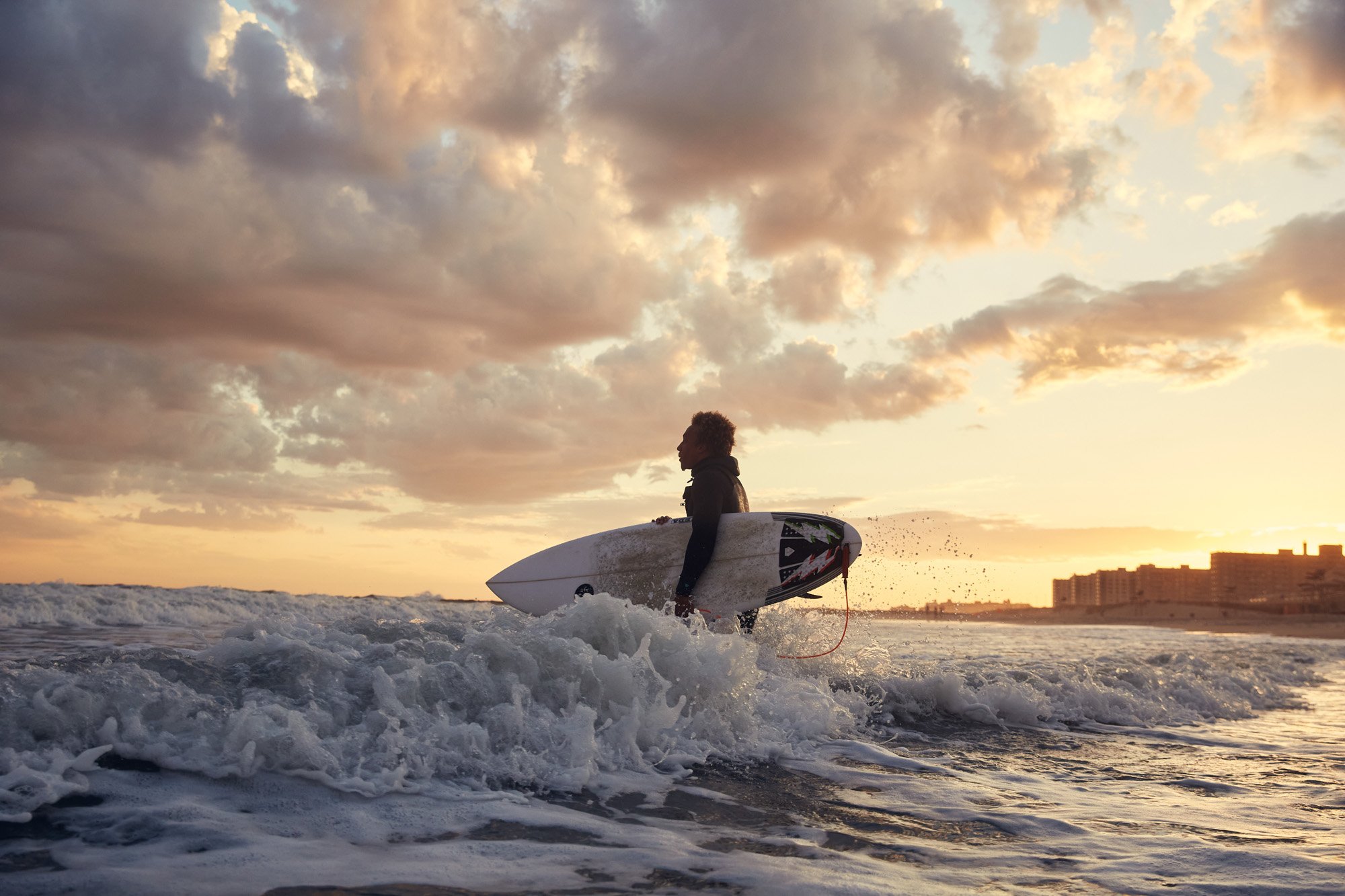 A surfer holding a surfboard walking in the ocean at sunset at Rockaway Beach New York City.