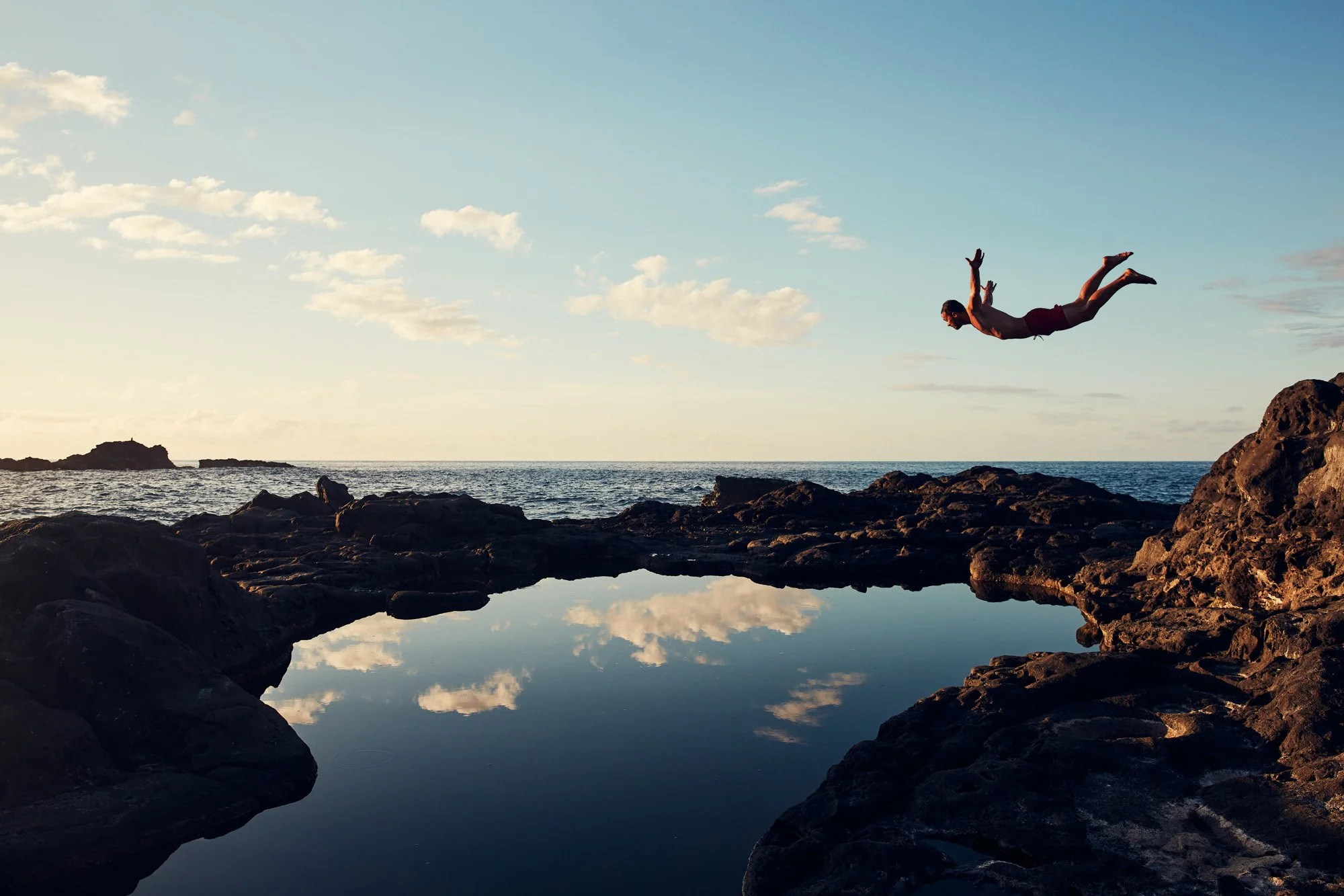 A person mid-air jumping off rocks into Olivine Pools on Maui Hawaii, reflecting the sky and clouds