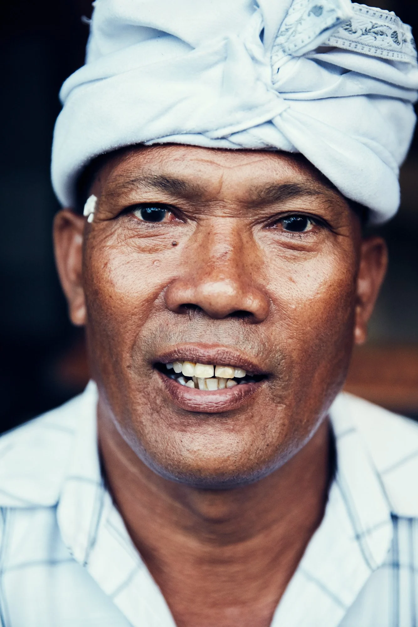 Close-up portrait of a Balinese man wearing a white turban with blue patterns, showing a warm expression.