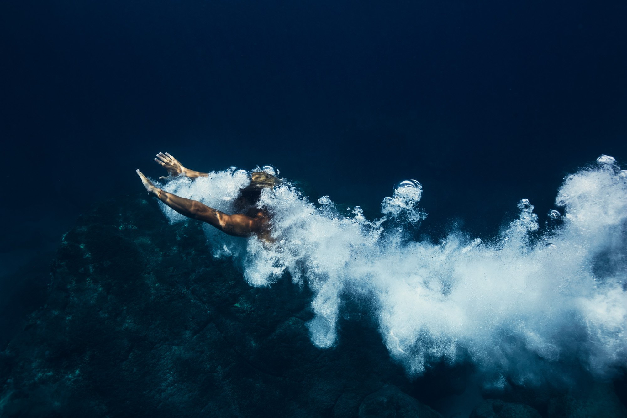 A person diving underwater, reaching forward with both arms, surrounded by splashing water and bubbles.