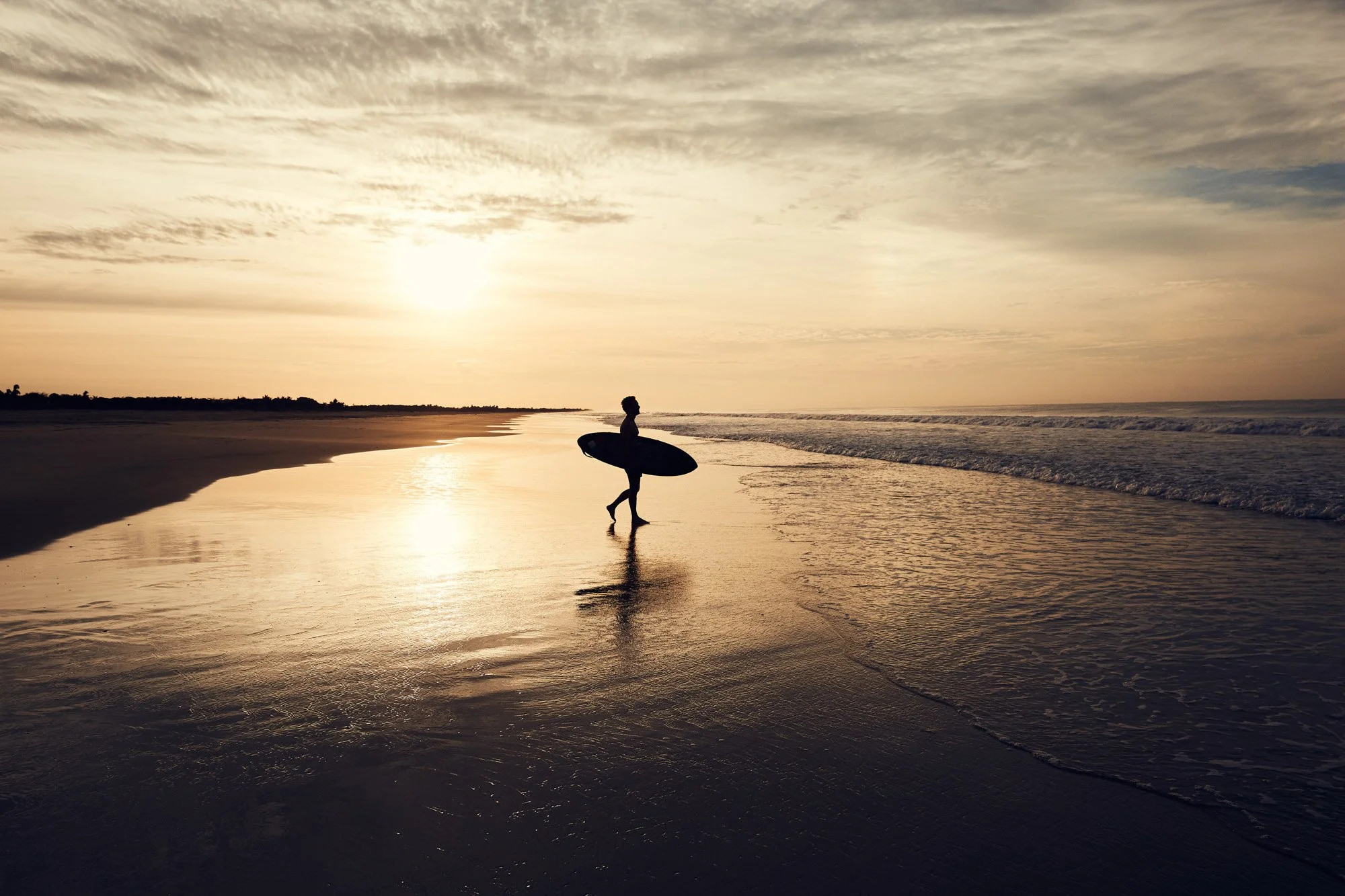 Person carrying a surfboard walking along the shoreline at sunrise on a quiet beach near Puerto Escondido Oaxaca Mexico.