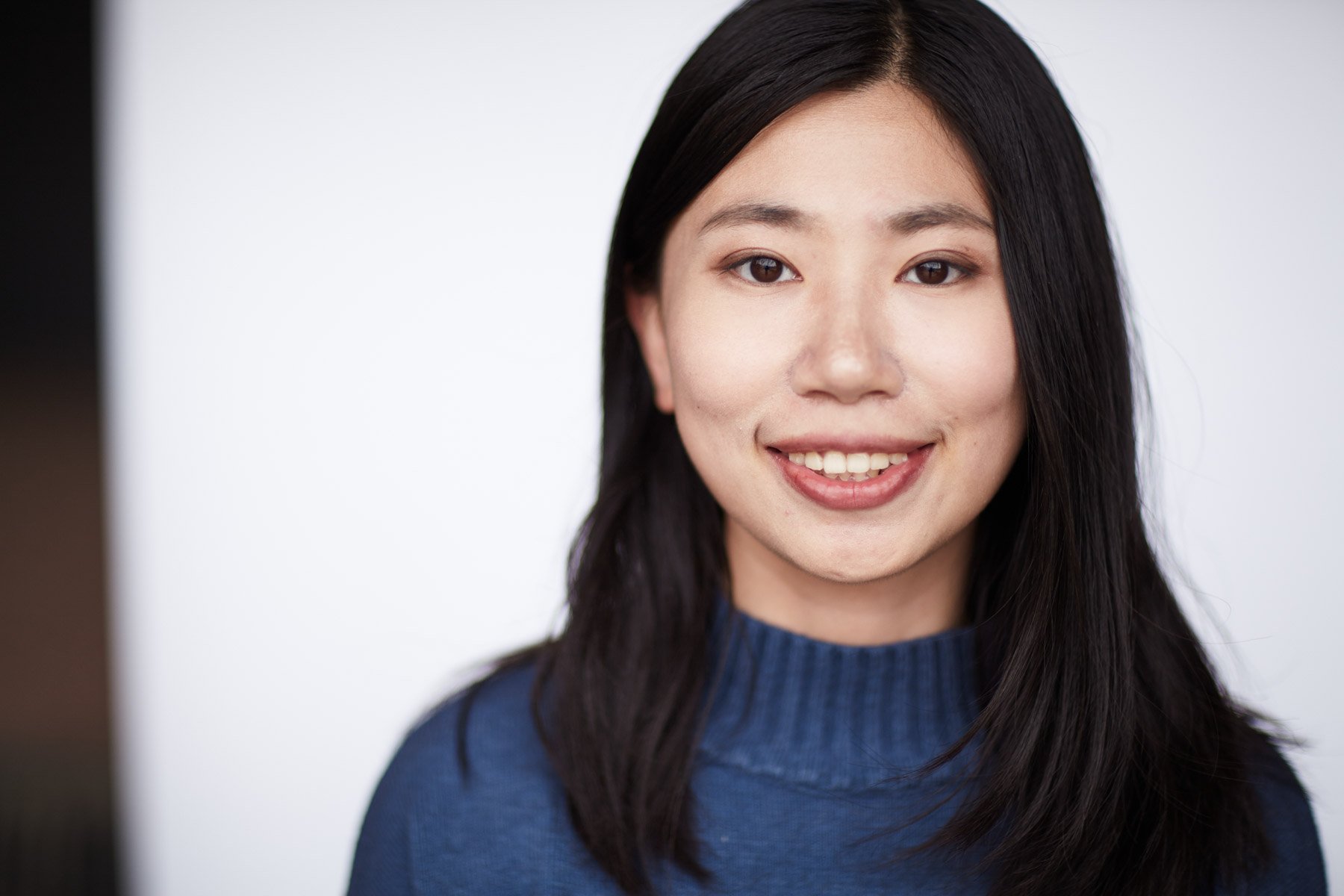 Portrait of a smiling Asian woman with long dark hair wearing a blue sweater, against a white background.