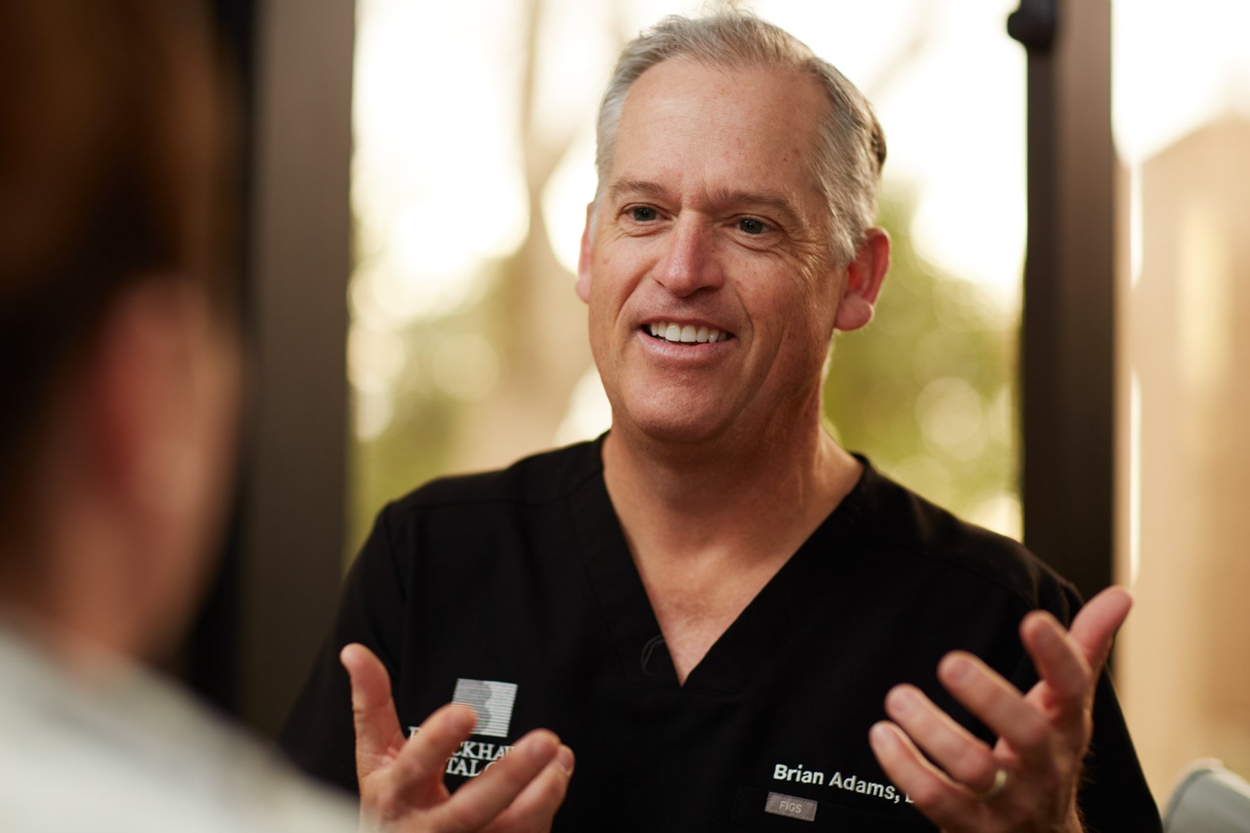 A smiling person in a black medical uniform, engaged in conversation, with blurred background.