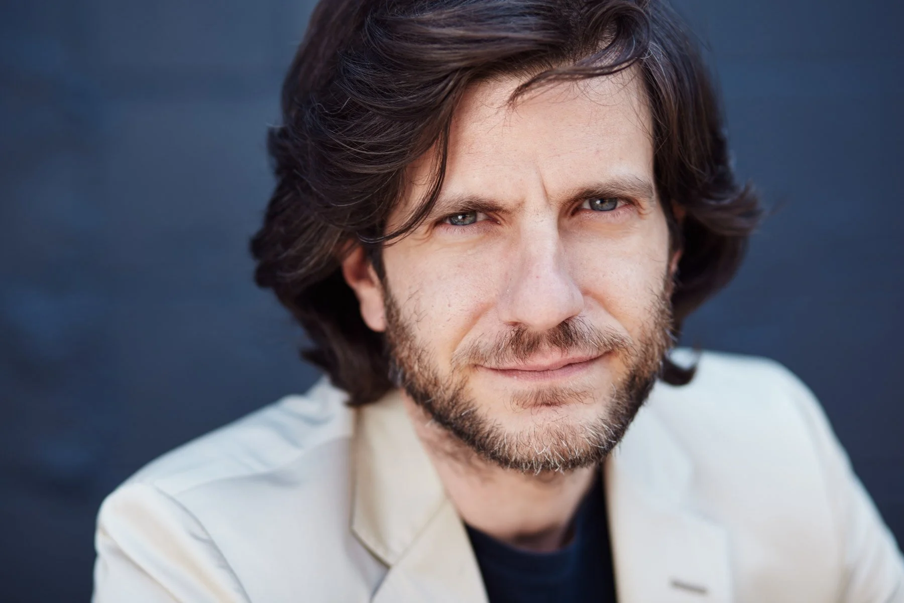 Close-up portrait of a man with long hair and a beard wearing a light-colored jacket, looking at the camera against a dark background, for Piaggio Fast Forward