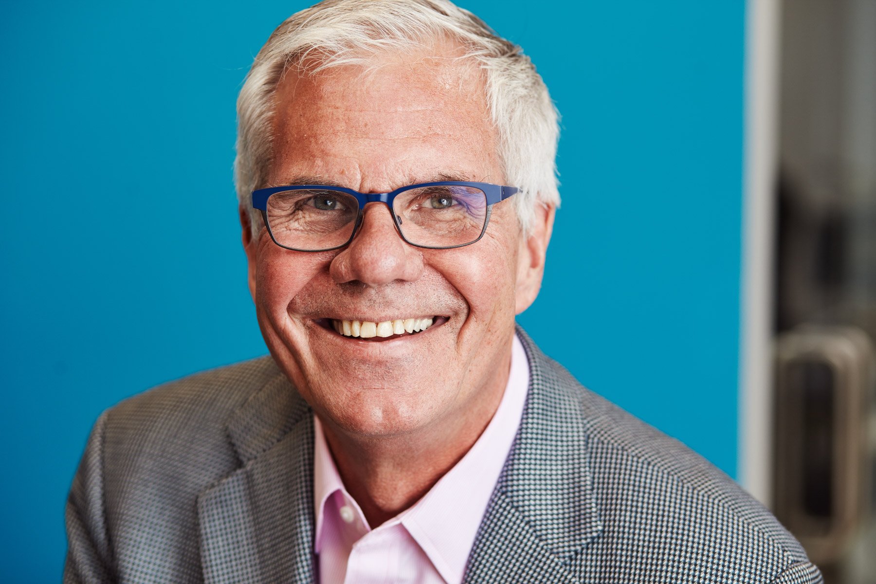 Smiling older man with glasses wearing a gray suit against a blue background for Piaggio Fast Forward in Boston Massachusetts