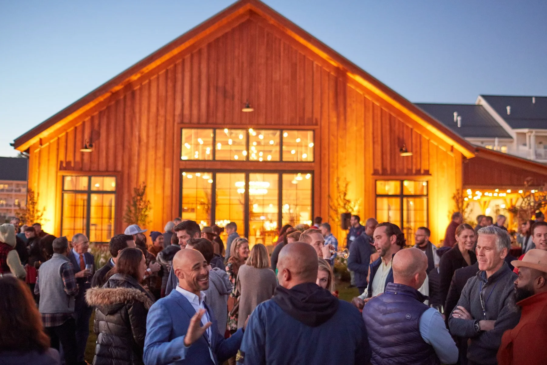 Outdoor evening gathering with people socializing in front of a warmly lit wooden building.