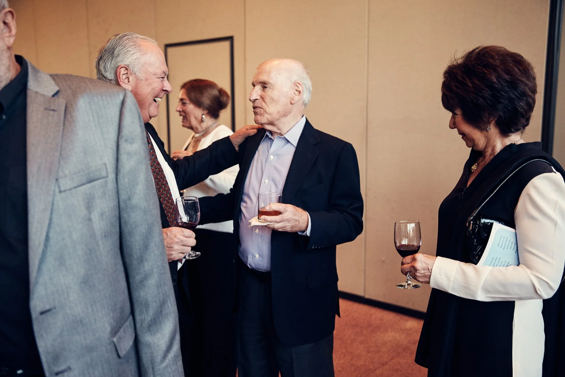 A small group of people socializing at an event, holding drinks and engaging in conversation. The setting appears formal, with attendees wearing suits and dresses.