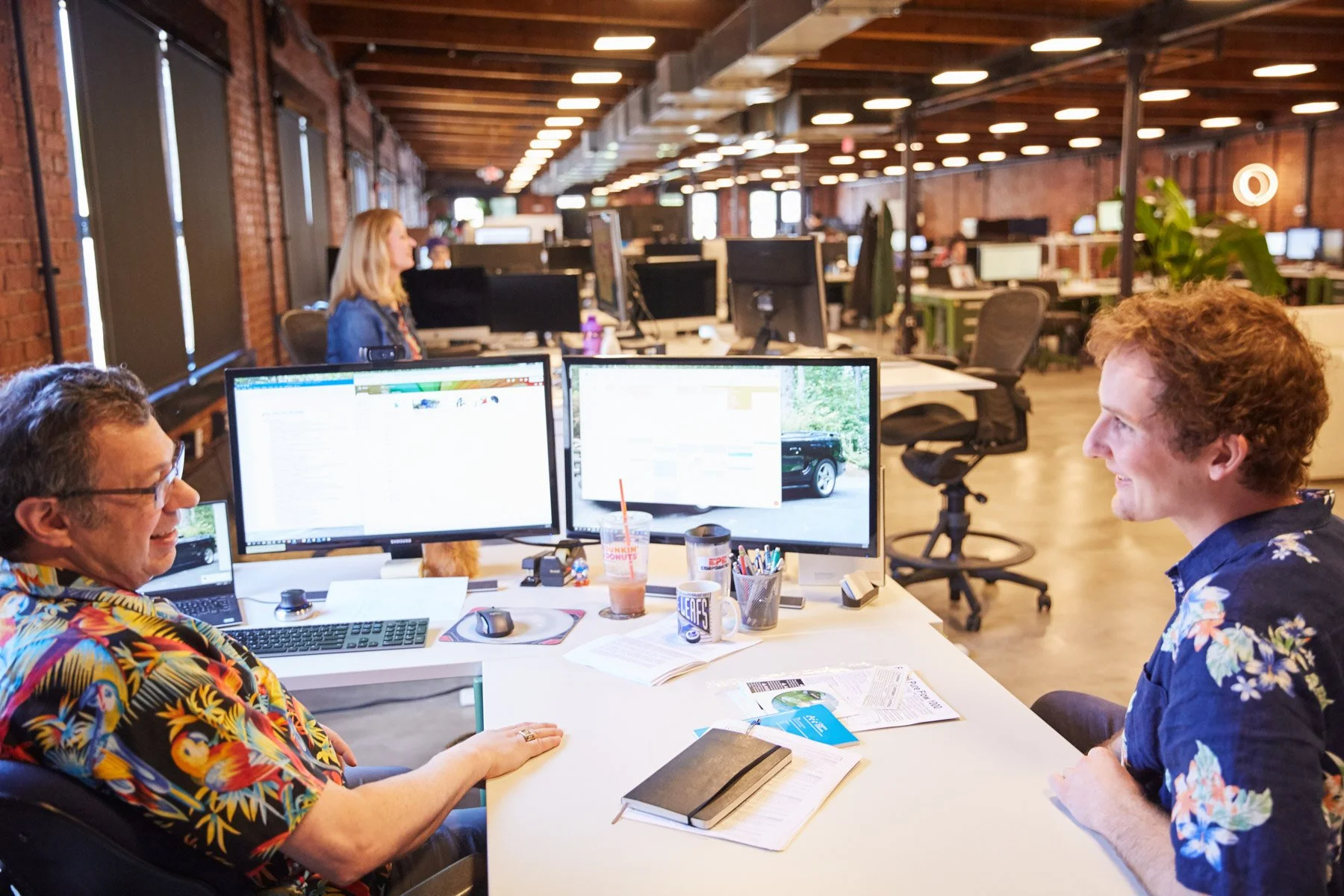 Two men sitting at a desk with computer monitors in an open office space, engaged in conversation. The office has exposed brick walls and ceiling lights at Piaggio Fast Forward in Boston Massachusetts
