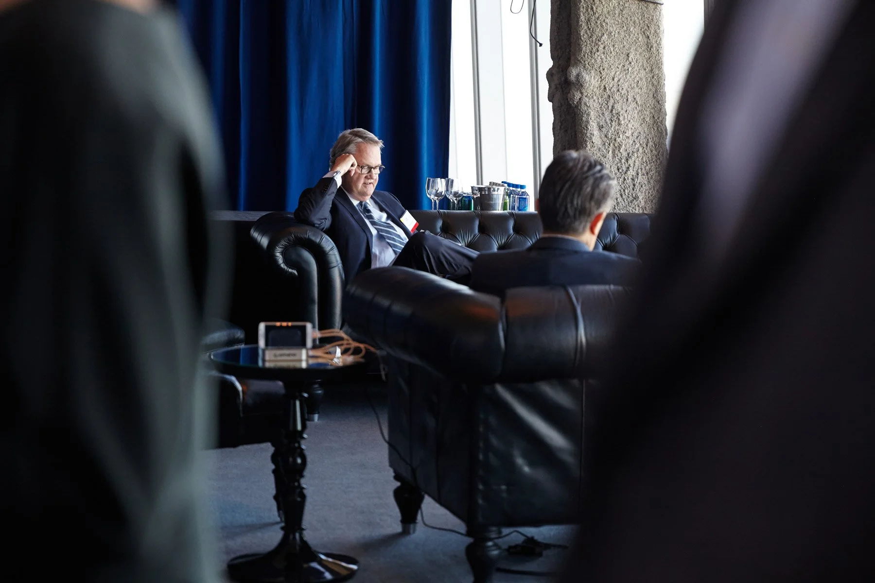 Two men seated on black leather couches in a modern, dimly lit lounge area.