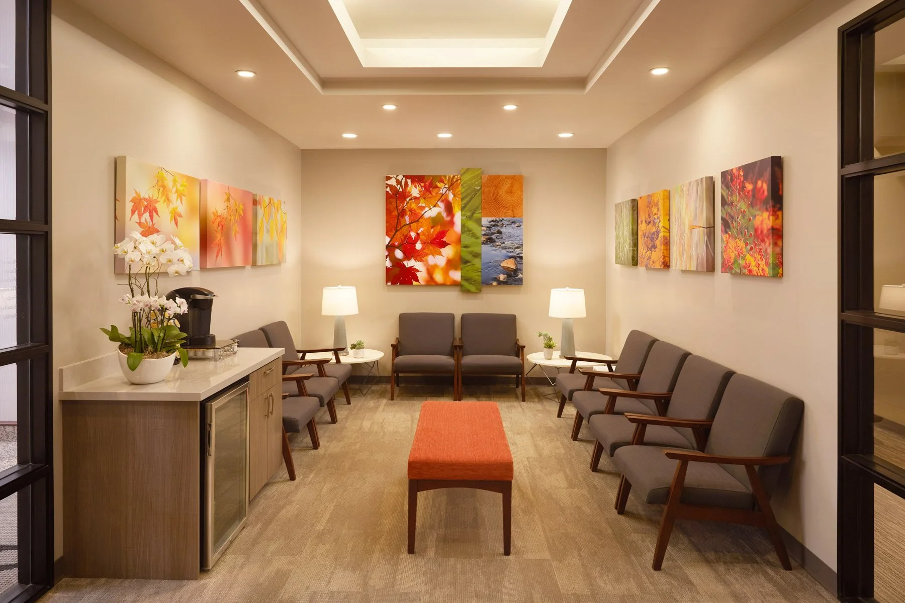 Modern waiting room with gray chairs, a red bench, abstract flower artwork on the walls, two lamps on side tables, a wooden cabinet with a coffee maker, and a potted orchid plant.