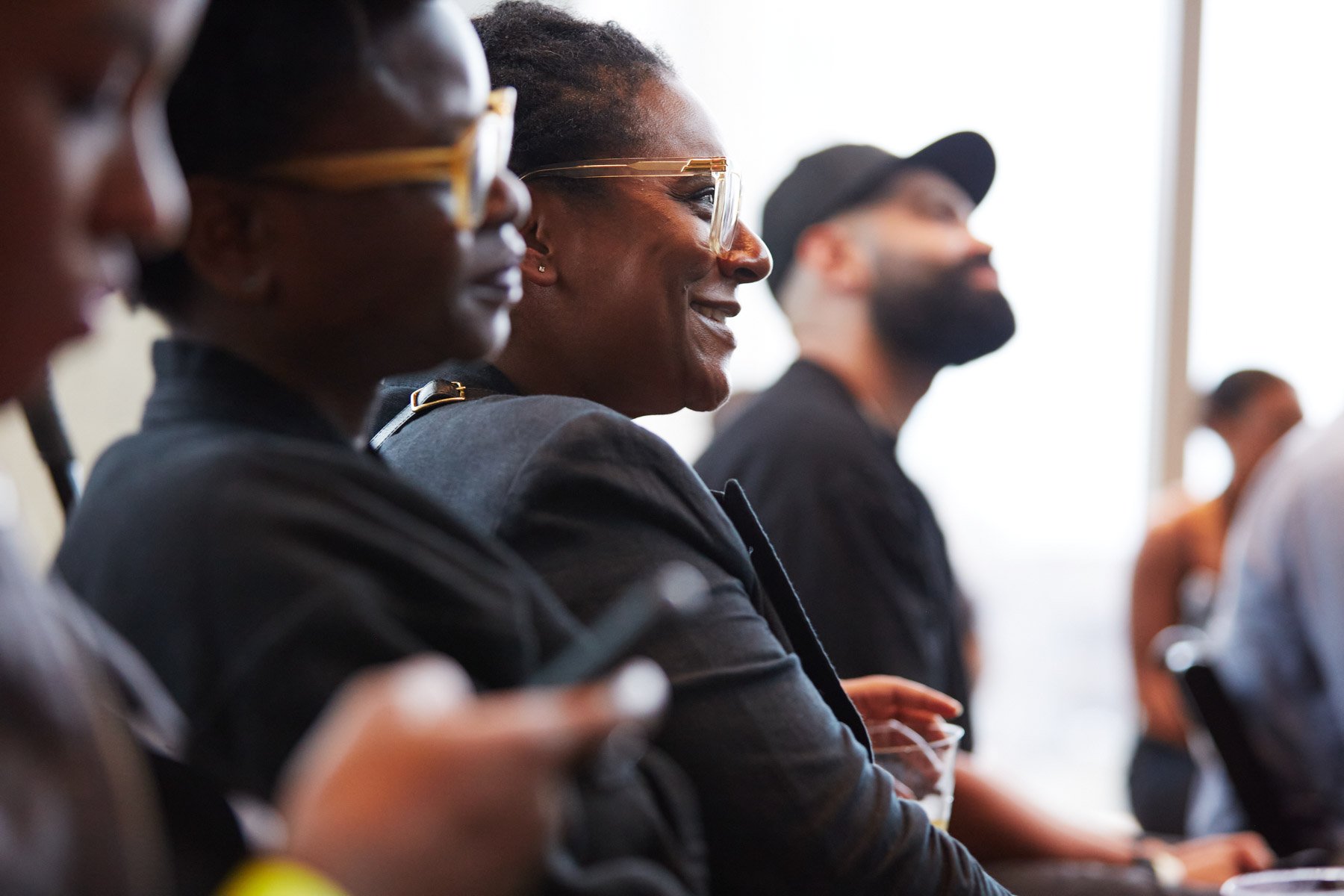 Audience members at an event sitting in chairs, with some using smartphones and one holding a drink, focused on a presentation off-camera at Tidal headquarters in Manhattan New York City