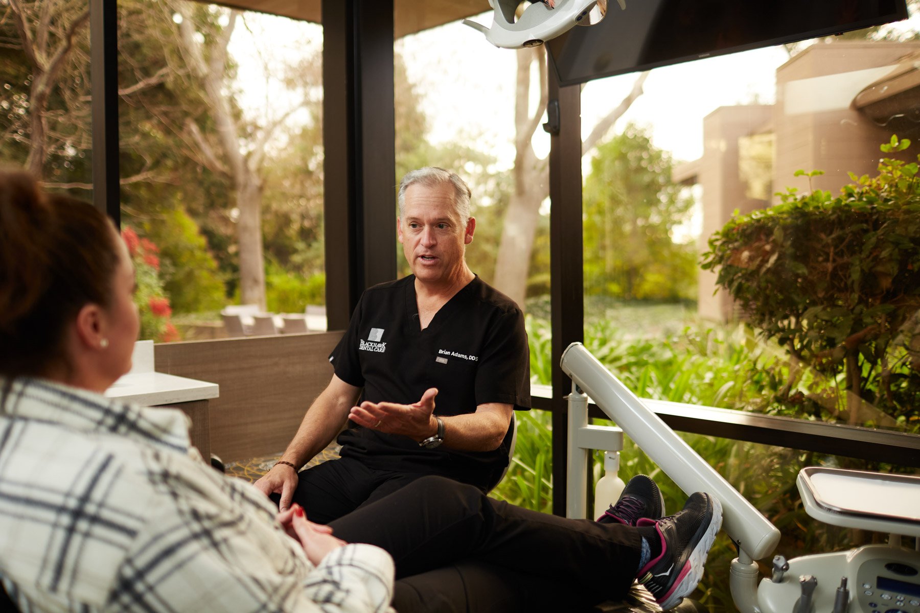 A dentist wearing a black uniform speaking to a patient in a dental office with large windows showing greenery outside.