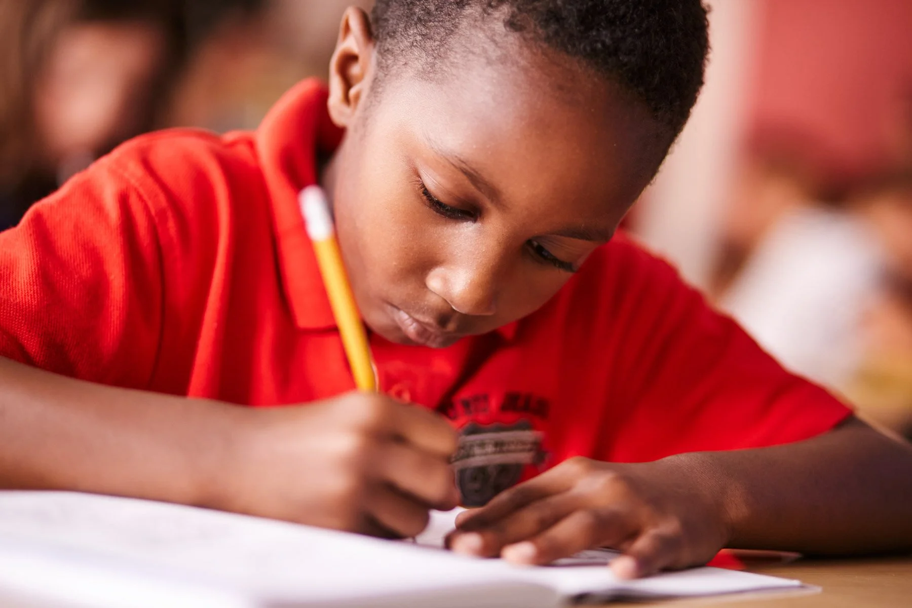 Young student in red shirt writing in a notebook with a pencil.