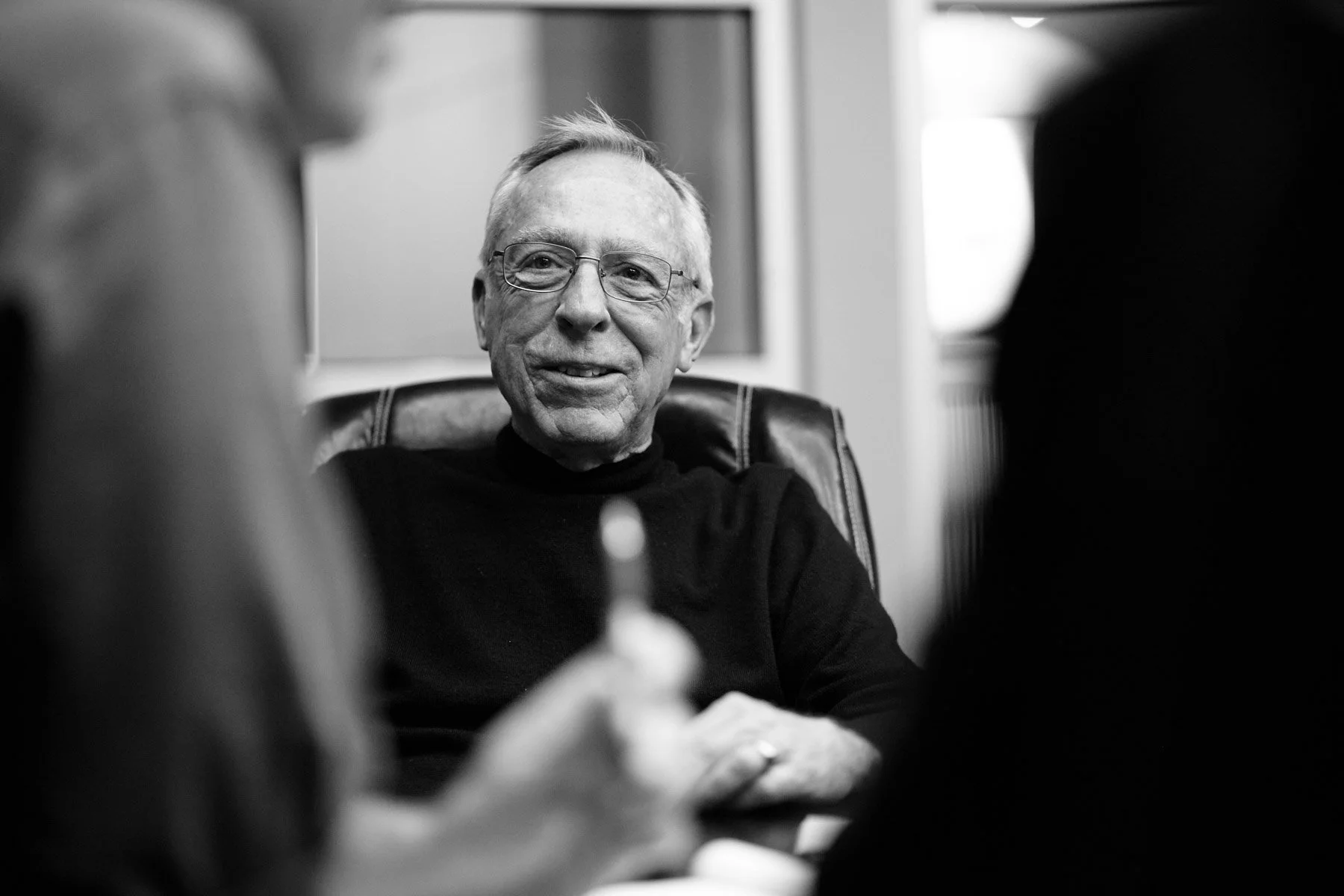 Black and white image of an elderly man wearing glasses and a black shirt, seated in an office chair, smiling, with blurred figures in the foreground for Wingman Advertising in Los Angeles California