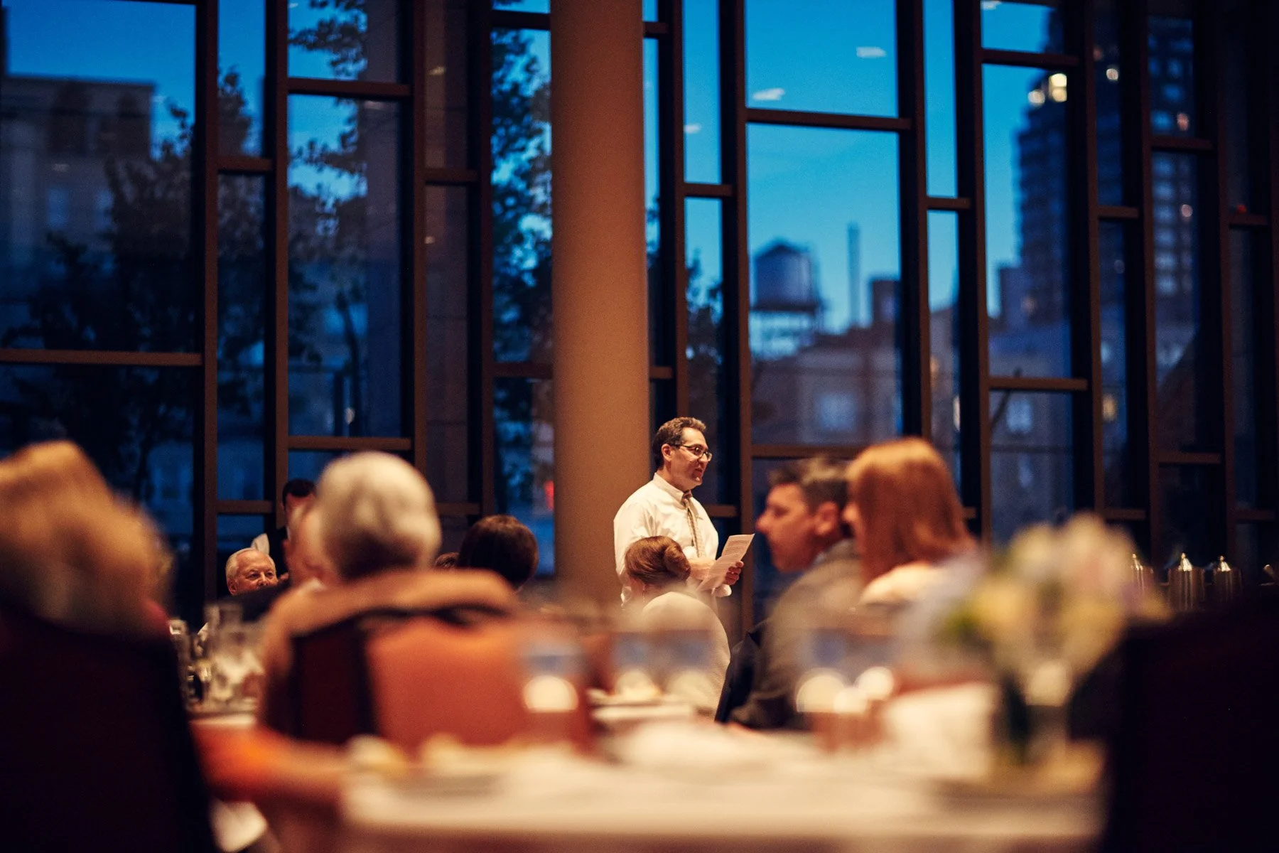 Man speaking in a restaurant or event venue, surrounded by seated guests with a cityscape visible through large windows.