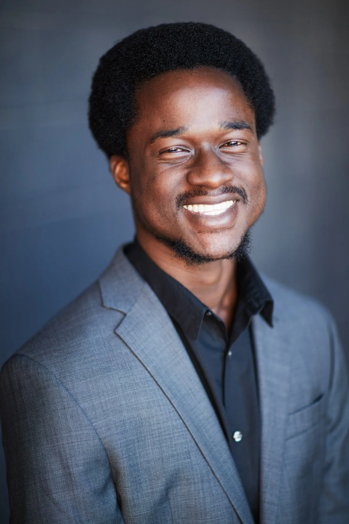 Man smiling in a gray suit and black shirt with a dark background.
