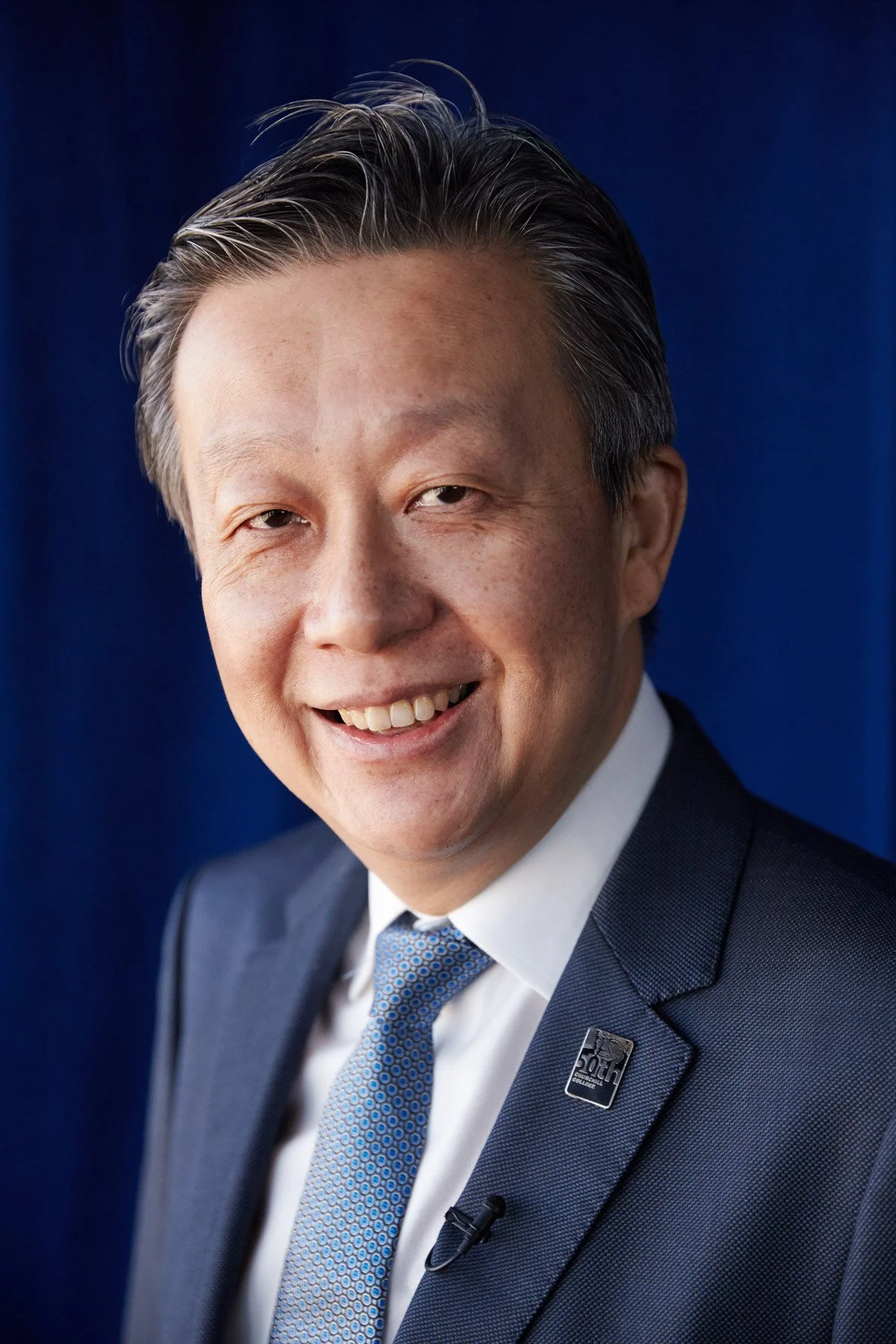 Smiling person in a suit with a tie, wearing a lapel pin, blue background.