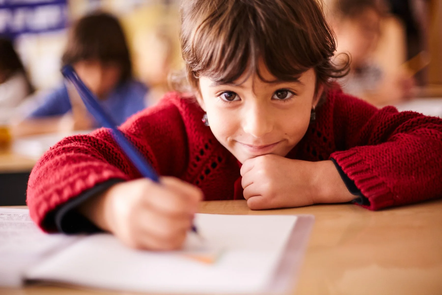 Young student in red sweater writing with a pen at a desk in a classroom, smiling at the camera.