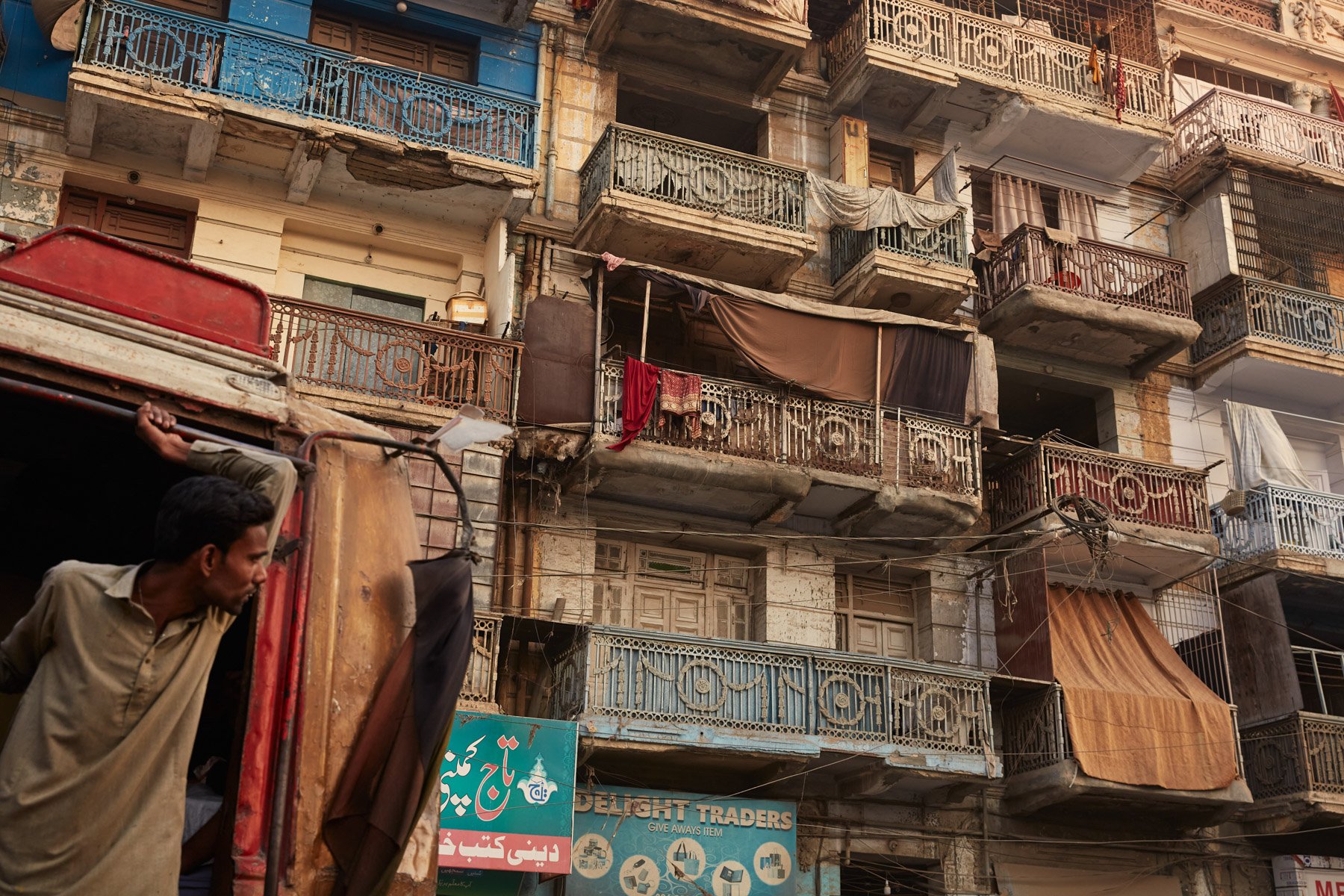 A multi-story apartment building with worn-out balconies, some with laundry hanging, and a man leaning out of a vehicle in the foreground near MA Jinnah Road in Karachi Pakistan.