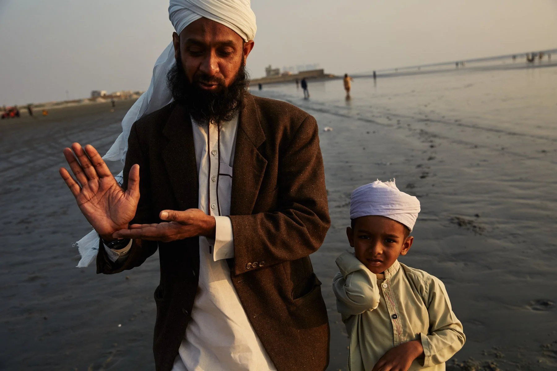 A man and a young boy wearing traditional Indian attire and white turbans on a beach with a bridge in the background. The man is gesturing with his hand and the boy looks curious or confused. Clifton Beach Karachi Pakistan