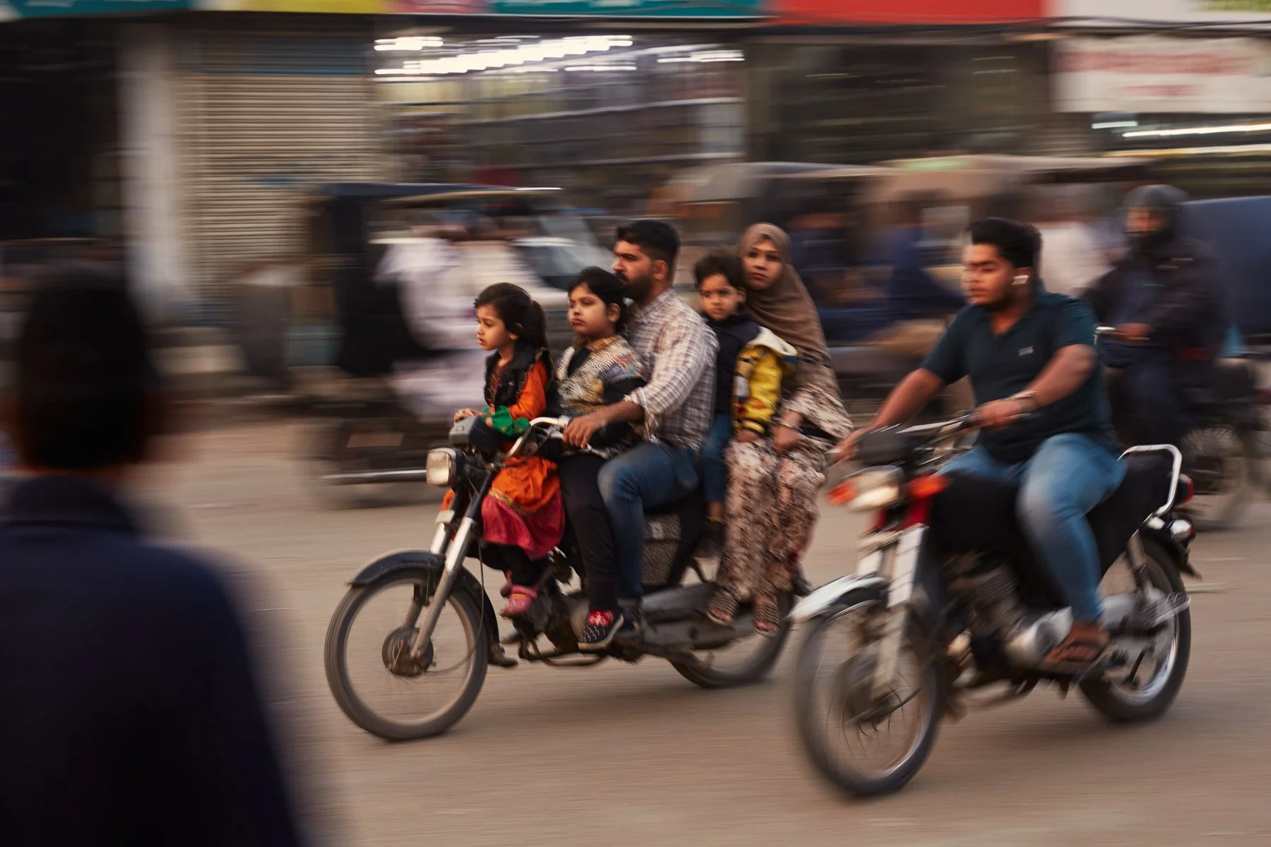 A family of six riding on a motorcycle on a busy street, with scooters and blurred vehicles in the background. Karachi Pakistan