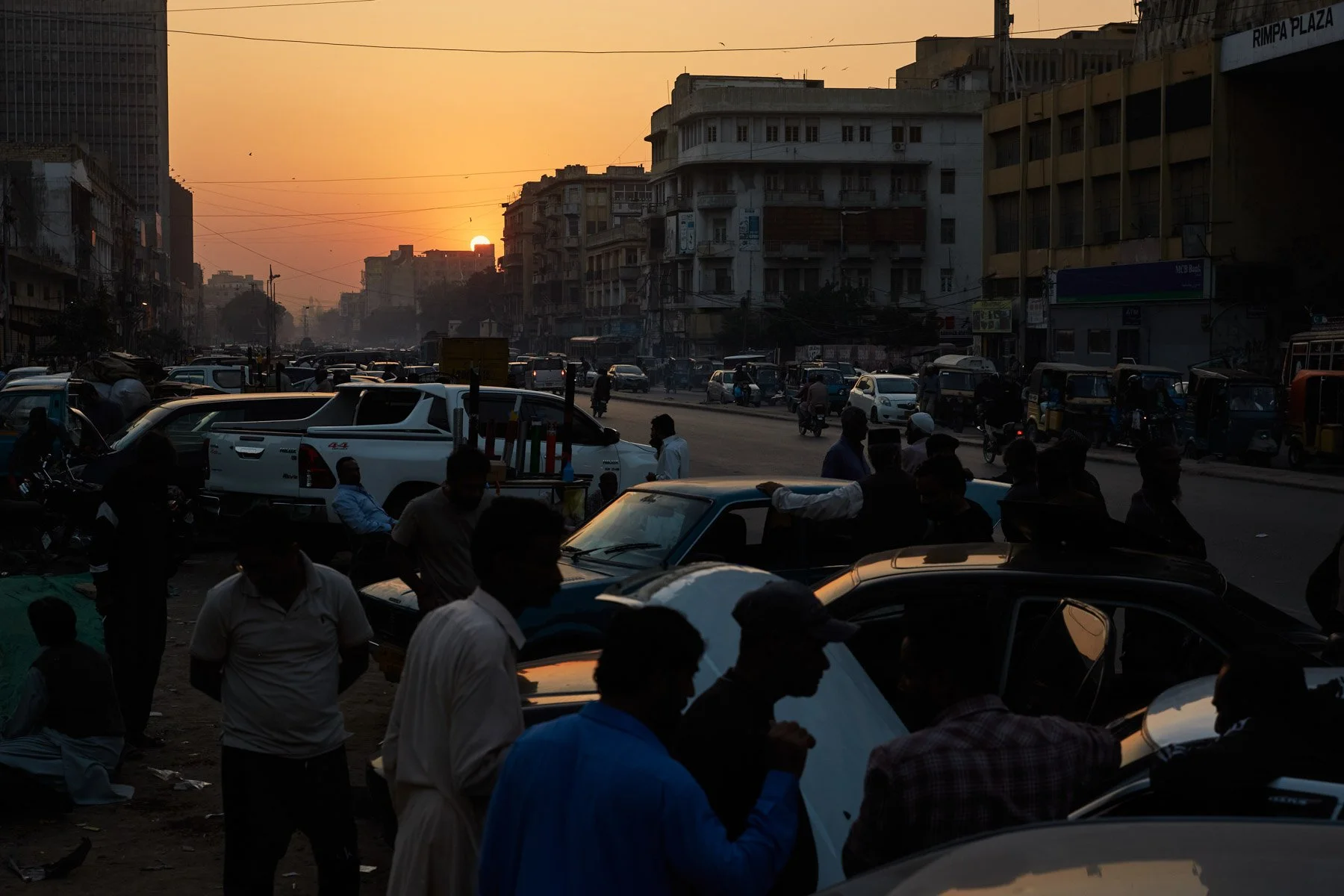MA Jinnah Road during sunset with cars, motorcycles, and pedestrians, silhouettes visible against the orange sky, buildings in the background. Karachi Pakistan