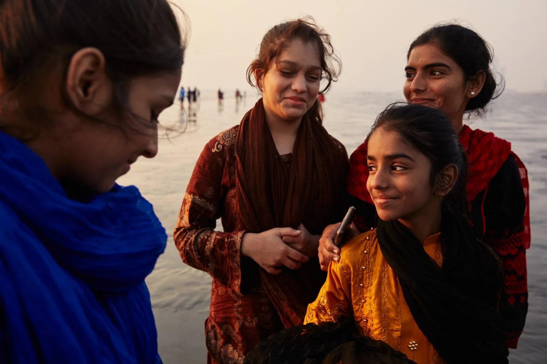 Group of four women standing by water during sunset, smiling, with one woman holding a smartphone. At Clifton Beach Karachi Pakistan
