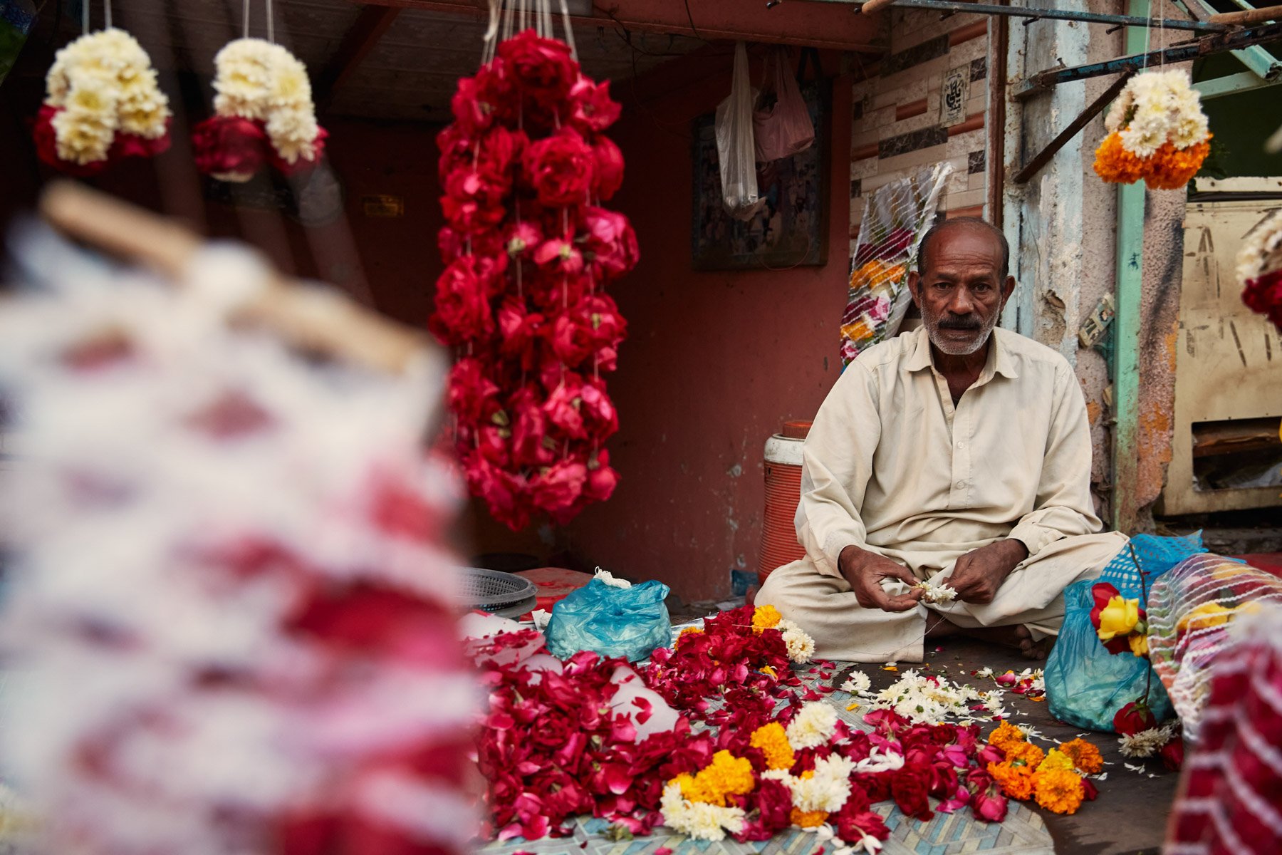 An elderly man sits on the ground surrounded by colorful flowers, including red, yellow, white, and orange, within a small shop or market stall with flowers hanging and scattered around. Near MA Jinnah Road in Karachi Pakistan