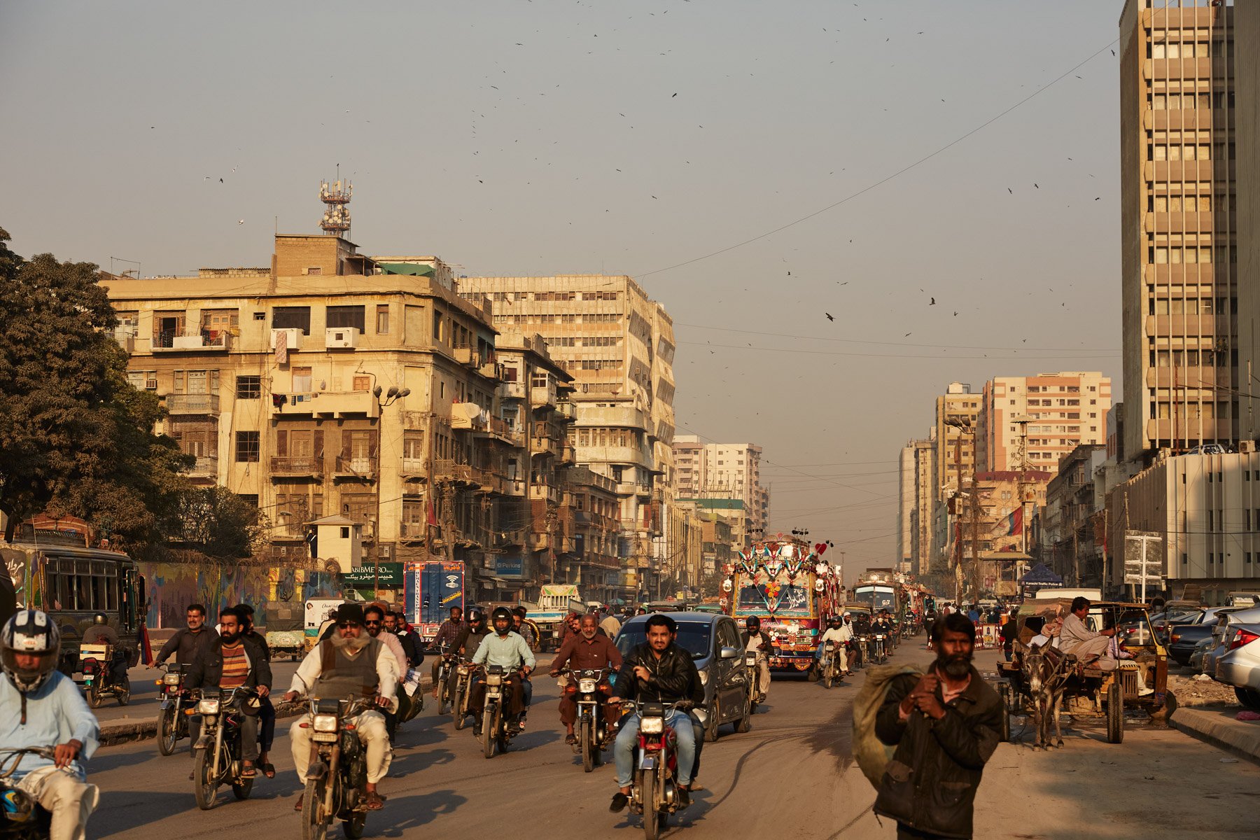 MA Jinnah Road with traffic including motorcycles, cars, and a decorative truck, surrounded by old and modern buildings, with many birds flying in the sky. Karachi Pakistan