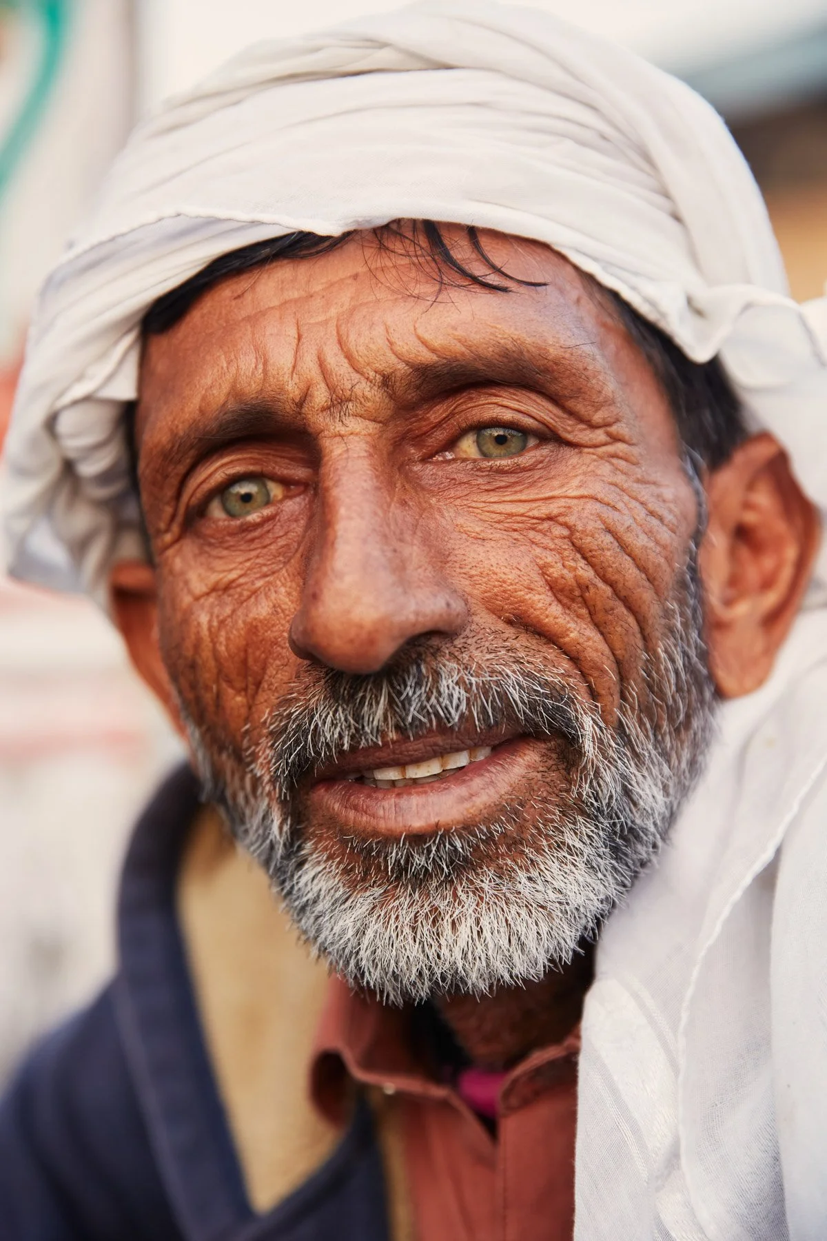 Close-up of an elderly fisherman with tan skin, gray beard, green eyes, wearing a white headscarf and a dark jacket, smiling gently, in the Dockyard of Karachi Pakistan.