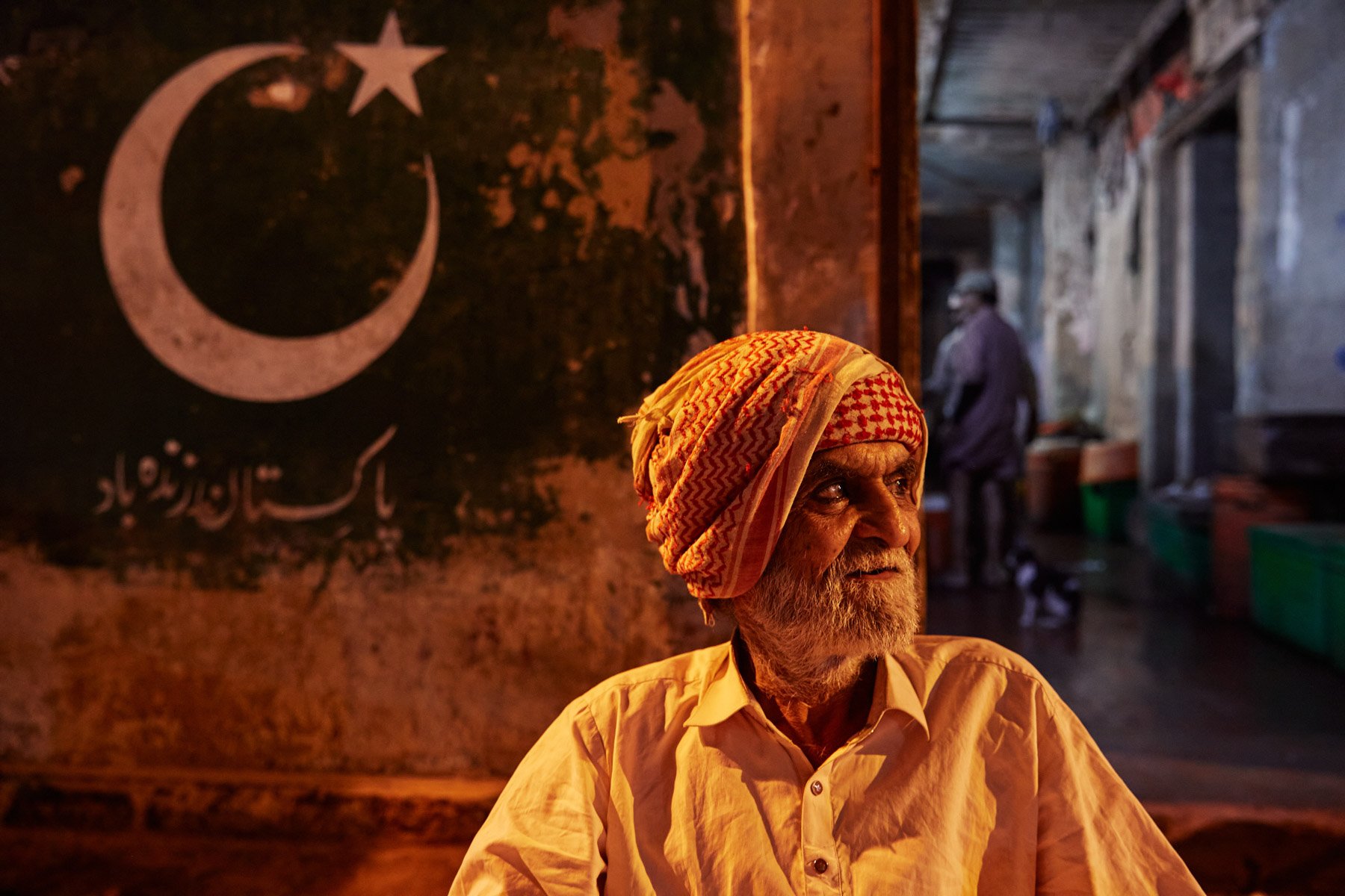 An elderly man with a white beard and a red patterned headscarf looks off to the side. Behind him, a dark wall displays a crescent moon, a star, and Arabic script, all illuminated by soft, warm lighting. Karachi Pakistan