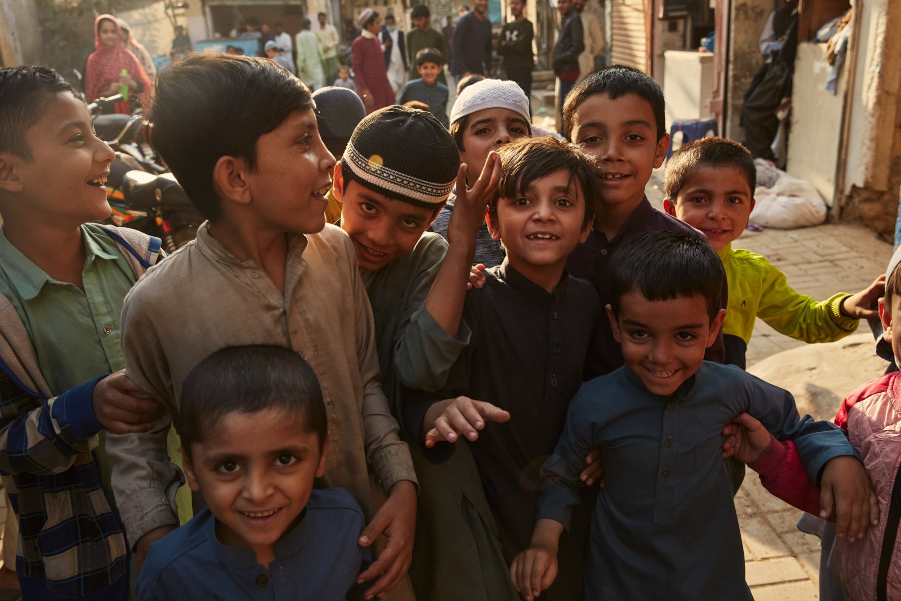 Group of smiling children gathered outdoors in a market or street setting, with some children wearing traditional clothing and others in casual attire, surrounded by market stalls and people in the background. Karachi Pakistan