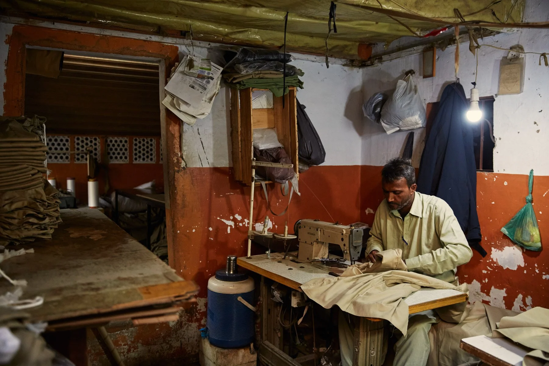 A man sewing fabric on a sewing machine in a small, cluttered workshop with a bare lightbulb hanging from the ceiling. Karachi Pakistan