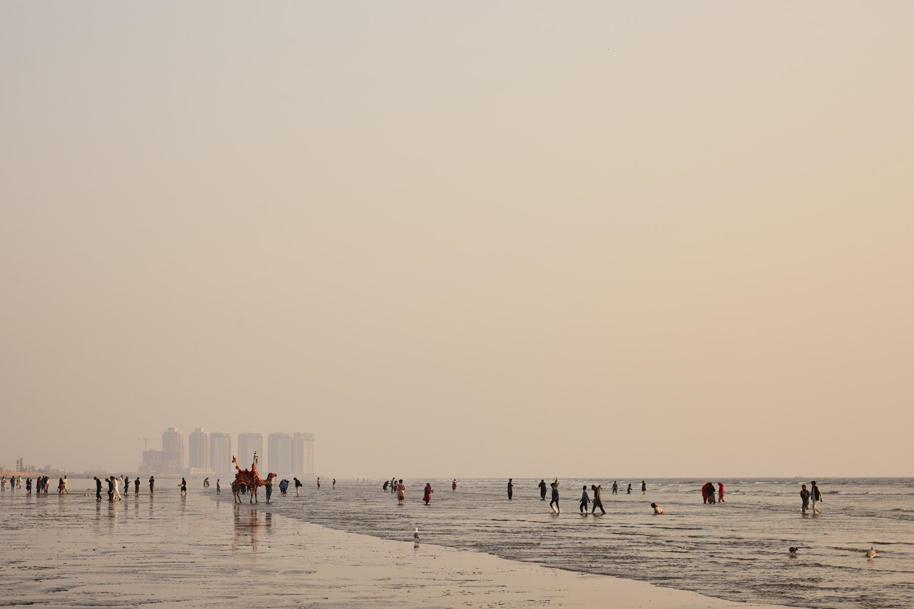 People walking on Clifton Beach, Karachi Pakistan with a camel in the foreground, high-rise buildings in the background, under a hazy sky.
