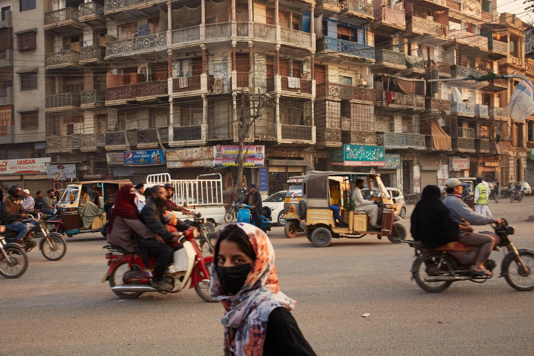Street busy with motorcycles, auto rickshaws, cars, and pedestrians in front of a worn multi-story building with balconies and shops in an urban area. MA Jinnah Road Karachi Pakistan
