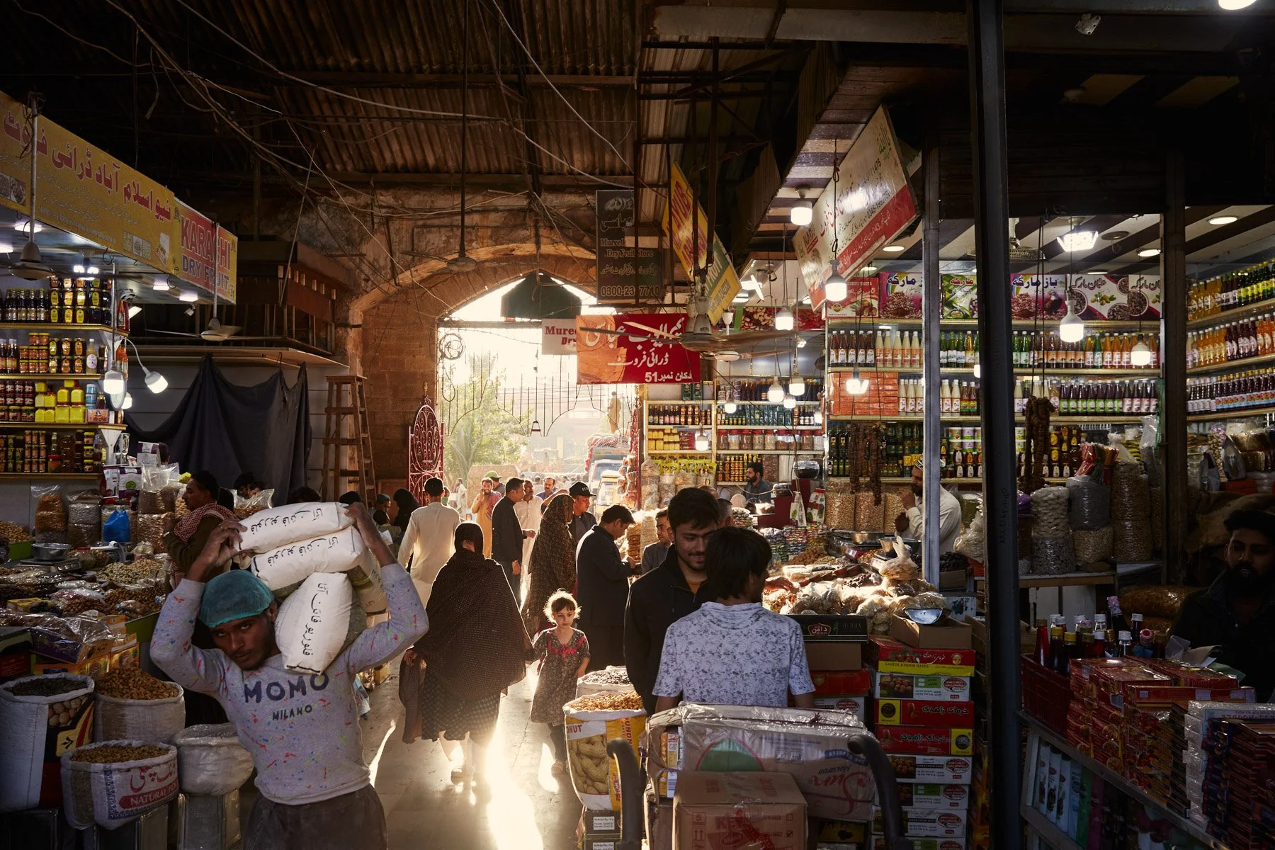 Empress Market with vendors and shoppers inside a partially open brick structure, shelves stocked with various goods, and sunlight streaming in from the entrance. Karachi Pakistan