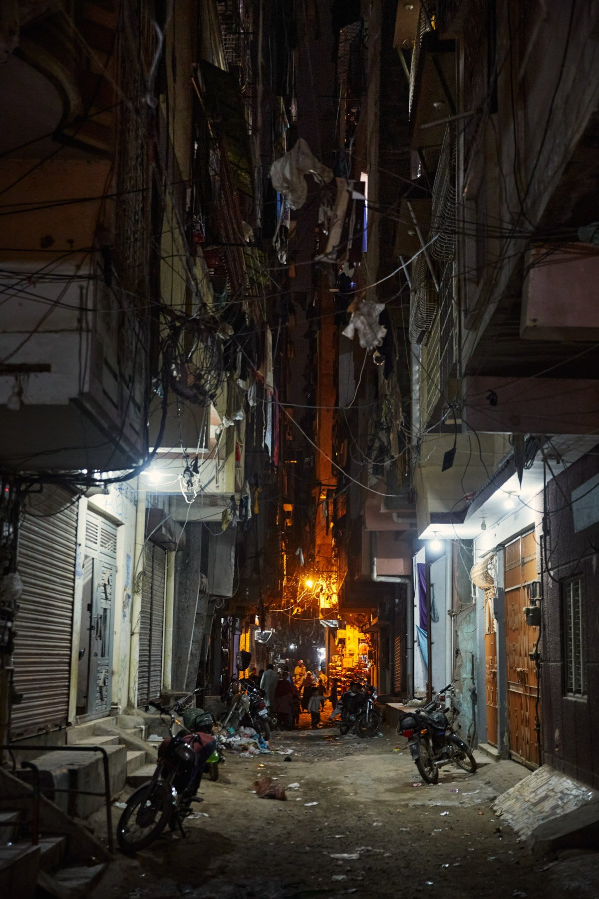 Nighttime scene of a narrow urban alley with buildings on both sides, showing cluttered electrical wires overhead, some motorcycles parked on the uneven dirt ground, and a few people in the distance near shops or street vendors. Karachi Pakistan