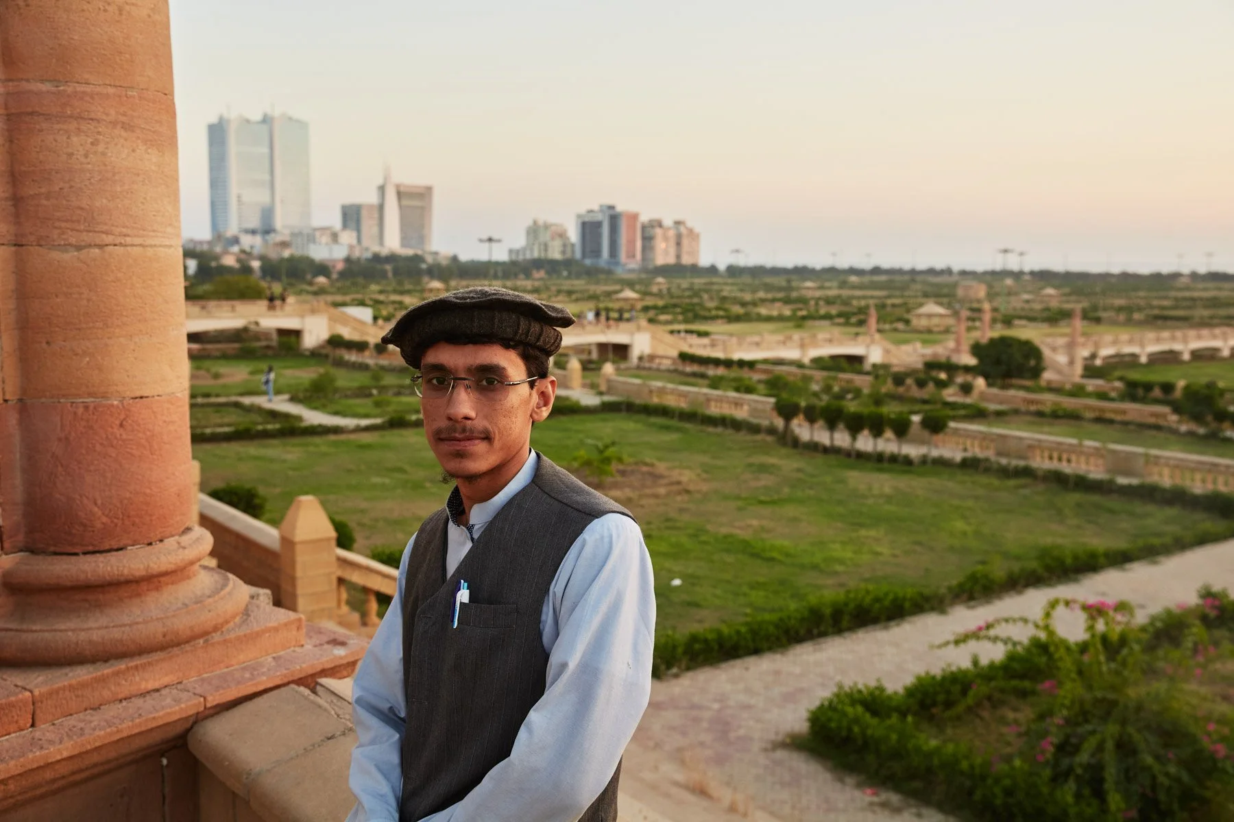 Young man in traditional attire standing in front of a historic monument with a cityscape of modern buildings in the background during sunset. Bagh Ibn-e-Qasim Park Karachi Pakistan