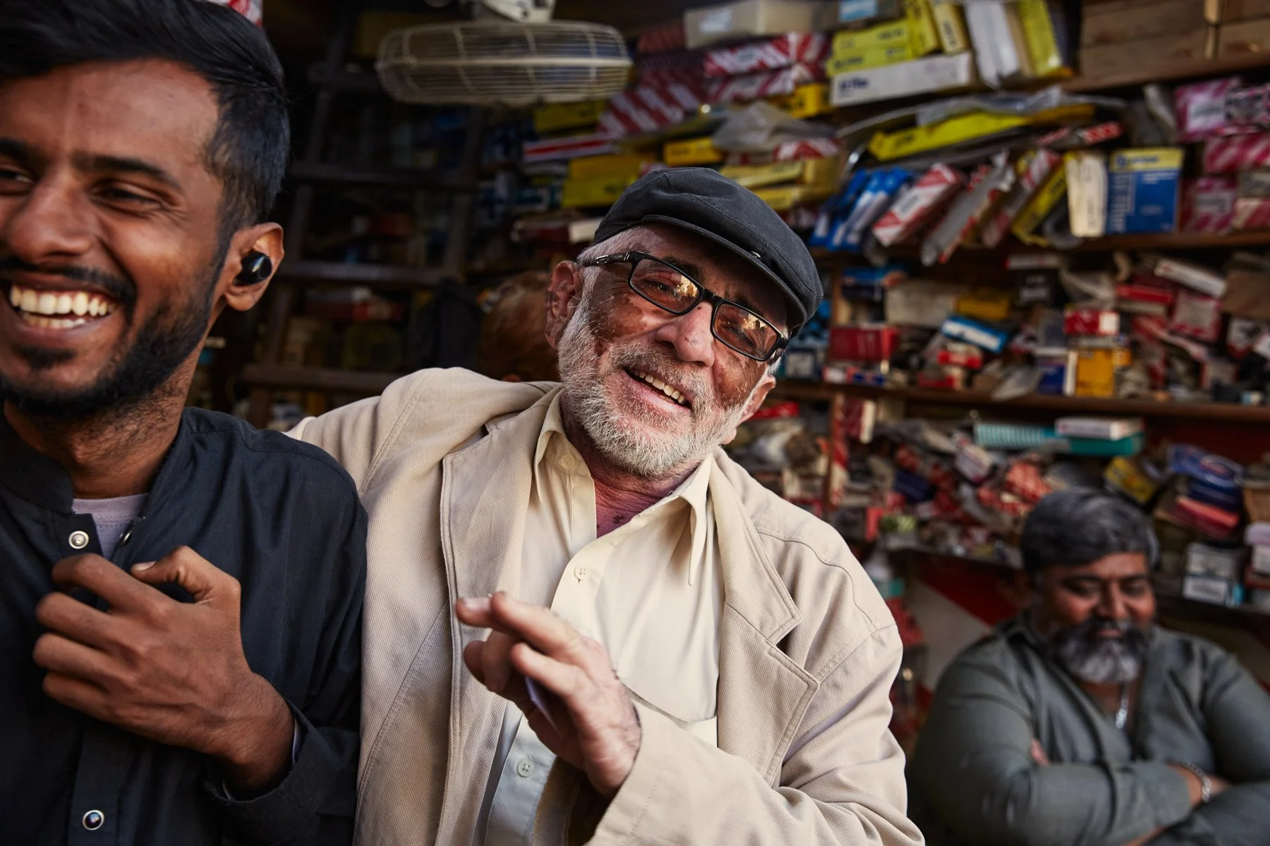 Three smiling men, one wearing a beige jacket and black cap, another in a black shirt, and a third in a gray shirt, sitting in front of shelves with various items. Near MA Jinnah Road Karachi Pakistan