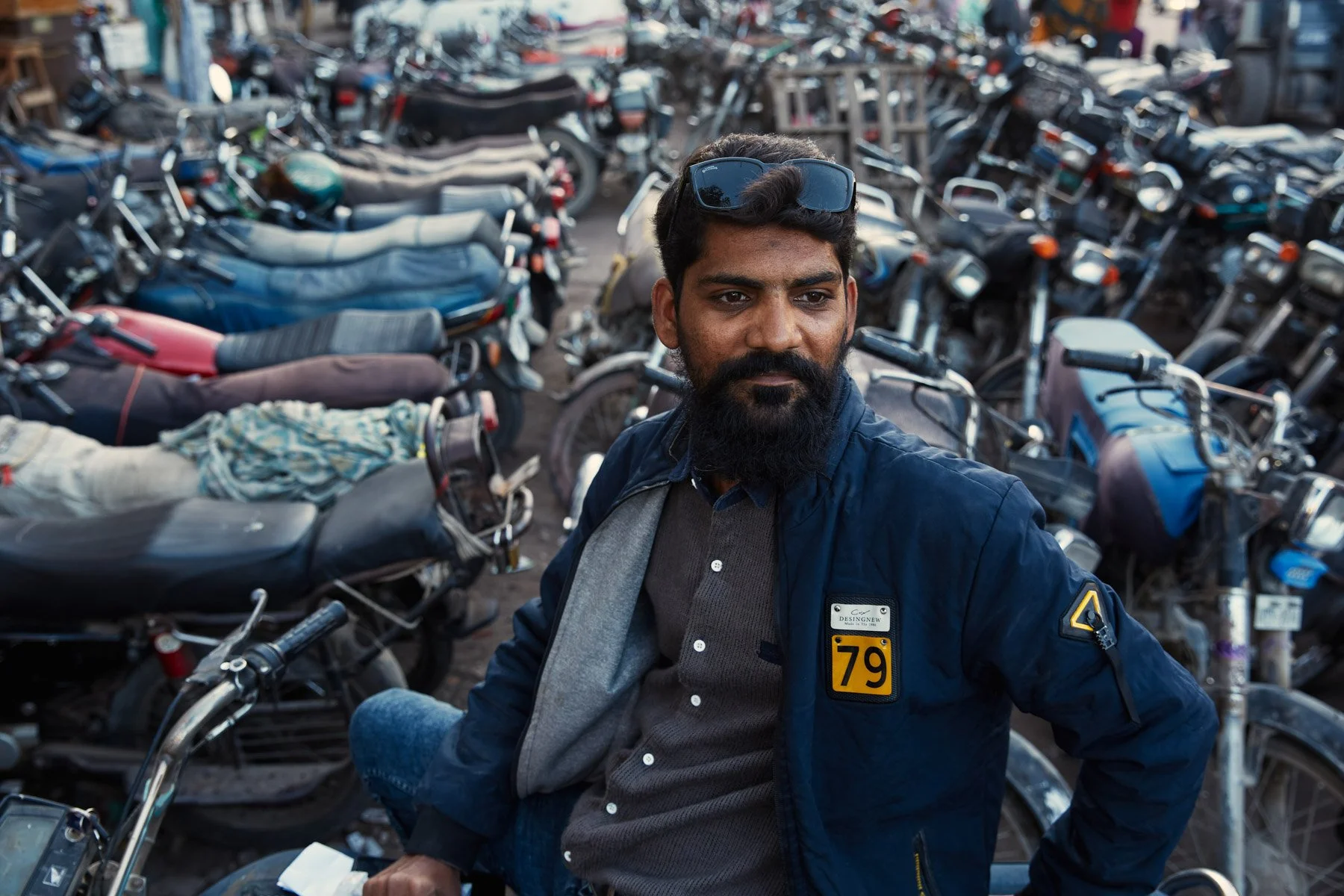 A man with dark hair and a beard sitting on a motorcycle, surrounded by rows of parked motorcycles in an outdoor lot in Karachi Pakistan