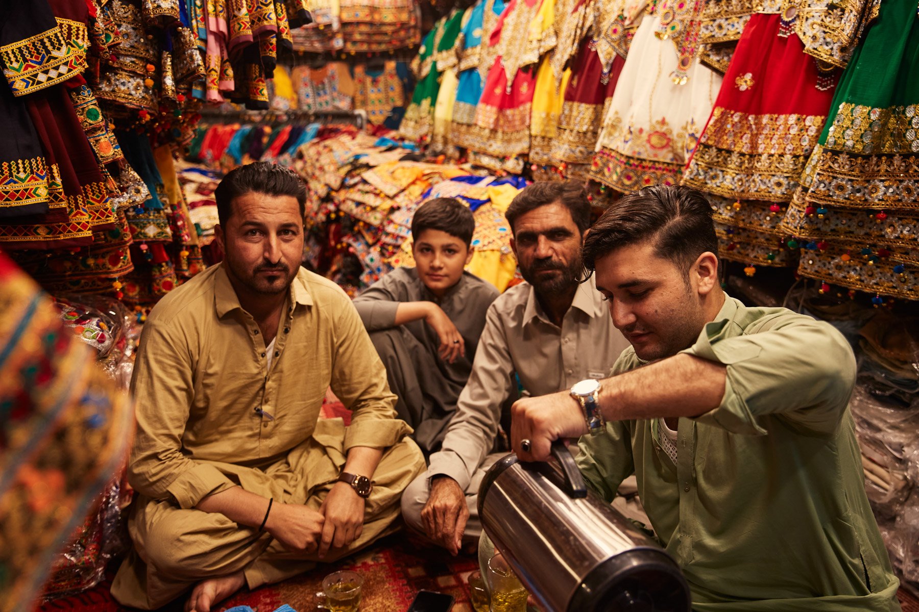 Four men and a boy sitting on the floor of a fabric stall filled with colorful embroidered clothes, with the man on the right pouring tea from a thermos. Karachi Pakistan