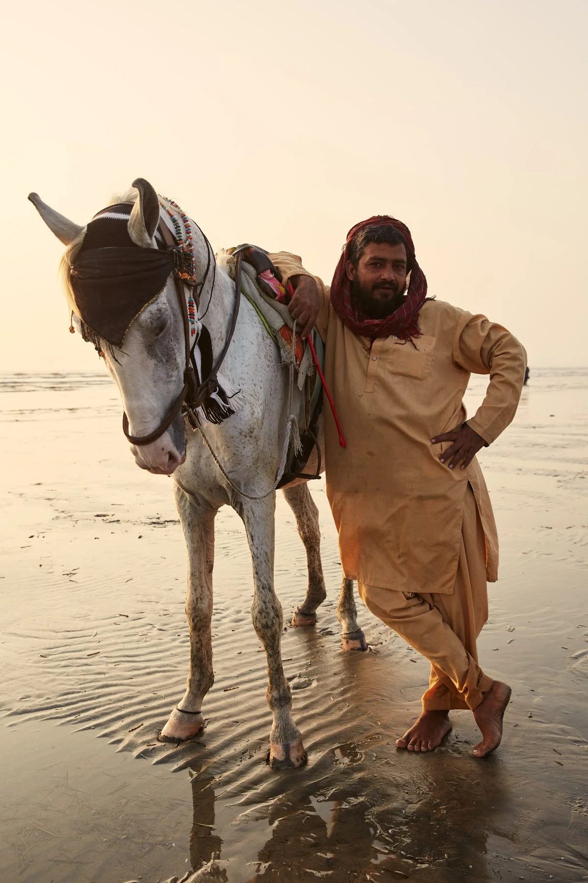 Man in traditional clothing standing with a decorated horse on a beach at sunset. Clifton Beach, Karachi Pakistan