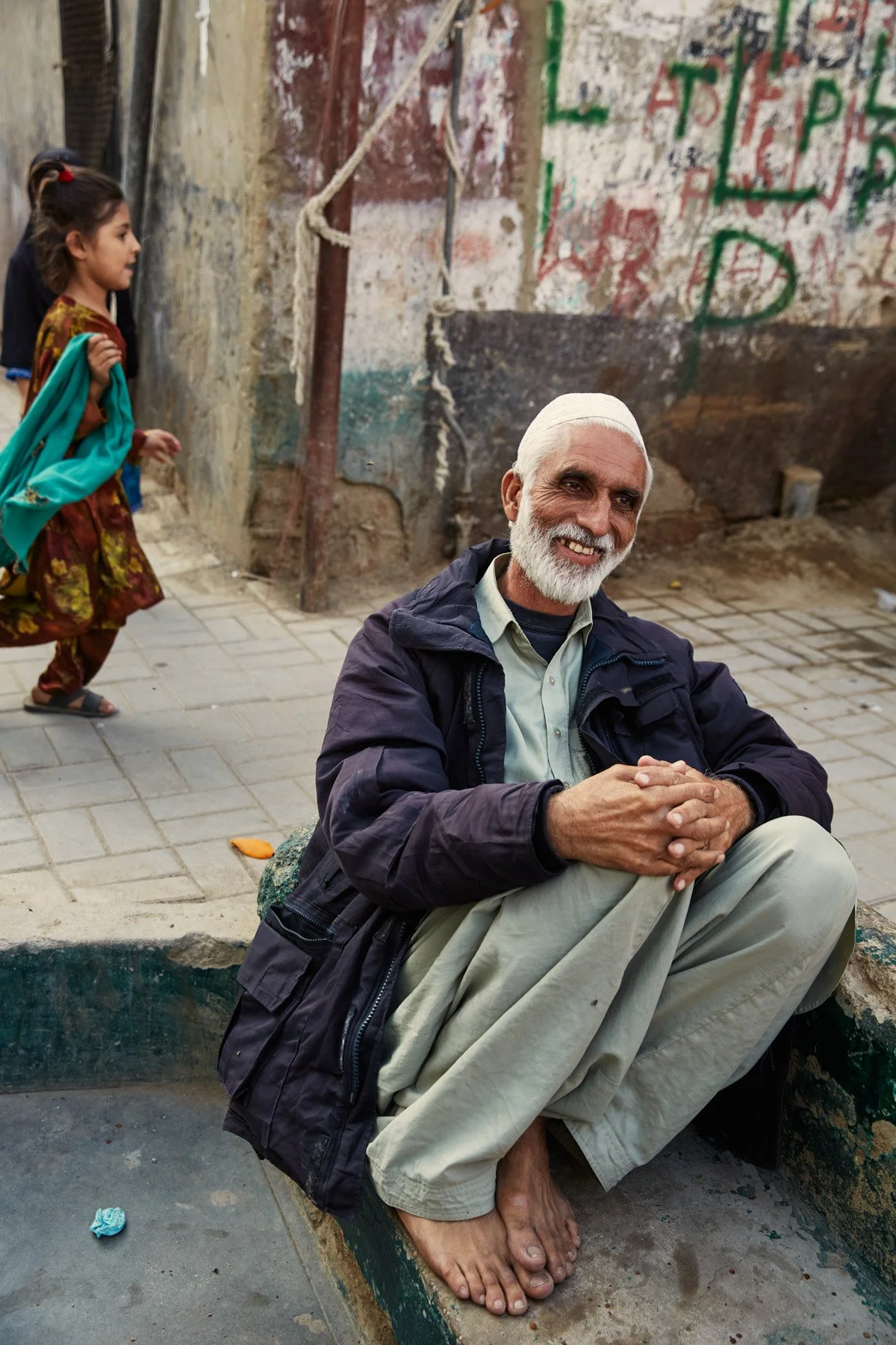 An elderly man with white hair and a beard smiling while sitting on a curb, with two young girls walking past in colorful traditional clothing in an outdoor, urban setting. Karachi Pakistan
