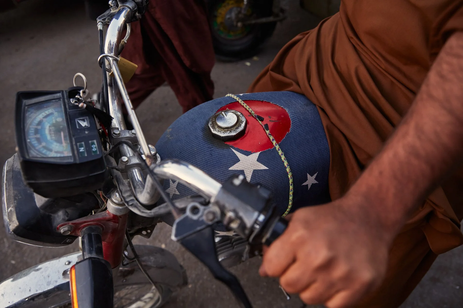 Close-up of a motorcycle's fuel tank with a weathered American flag and a chain, held by a person wearing brown clothing, in Karachi Pakistan.