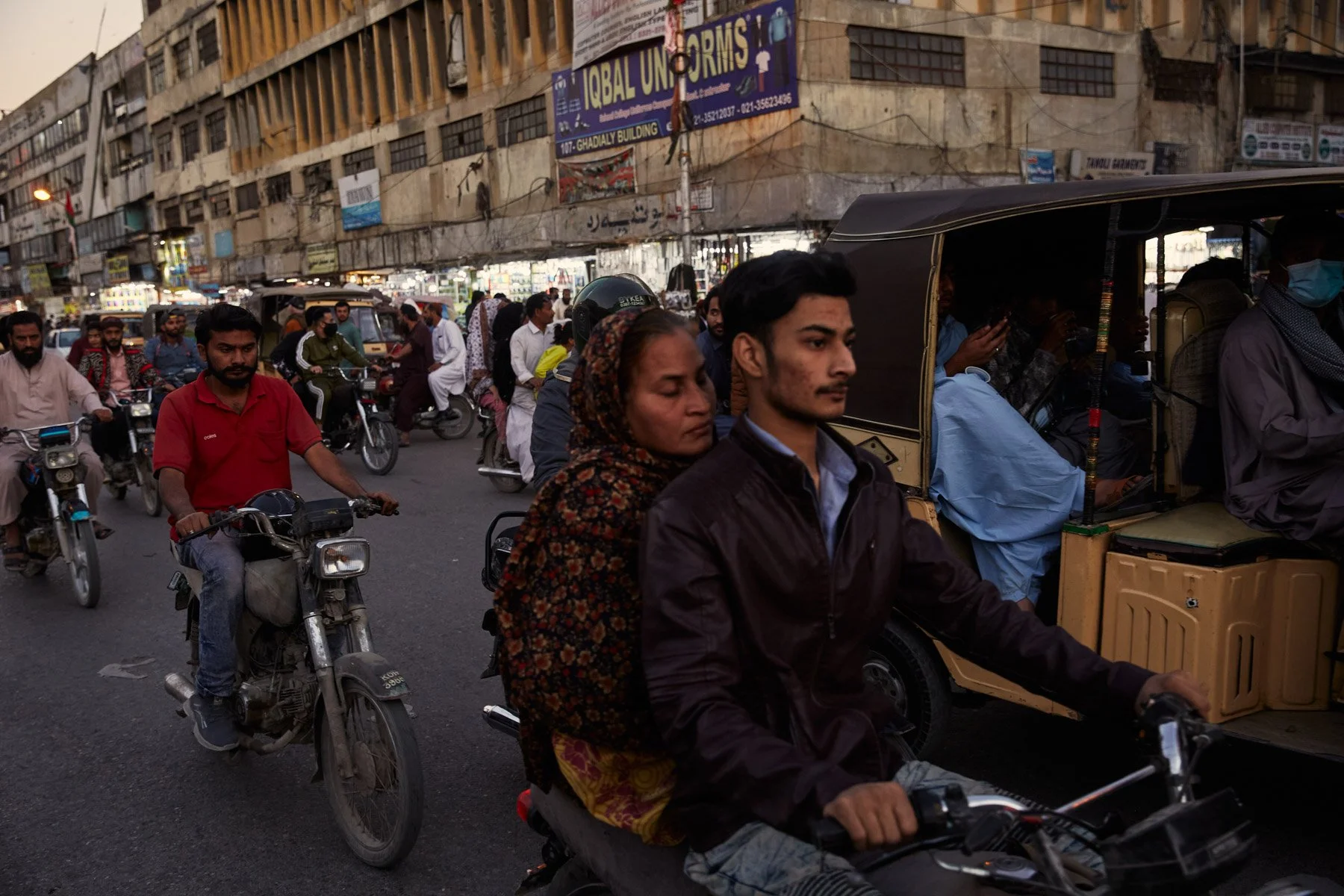 Street scene in a busy city with many people riding motorcycles and a rickshaw. The background shows a multi-story building with various signs and storefronts. Karachi Pakistan